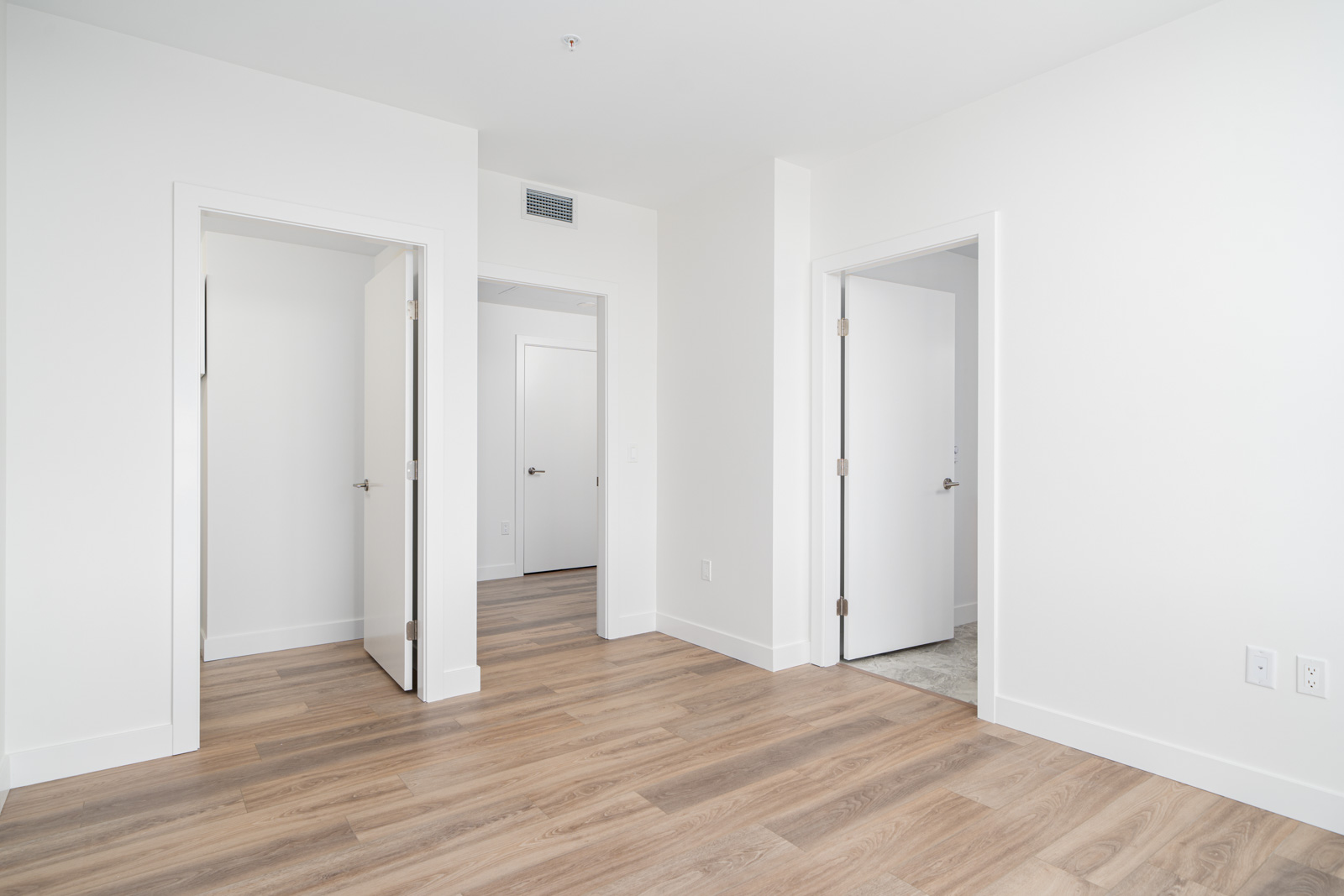 Empty room with light-colored walls, wood-patterned flooring, and three open white doors leading to other rooms.