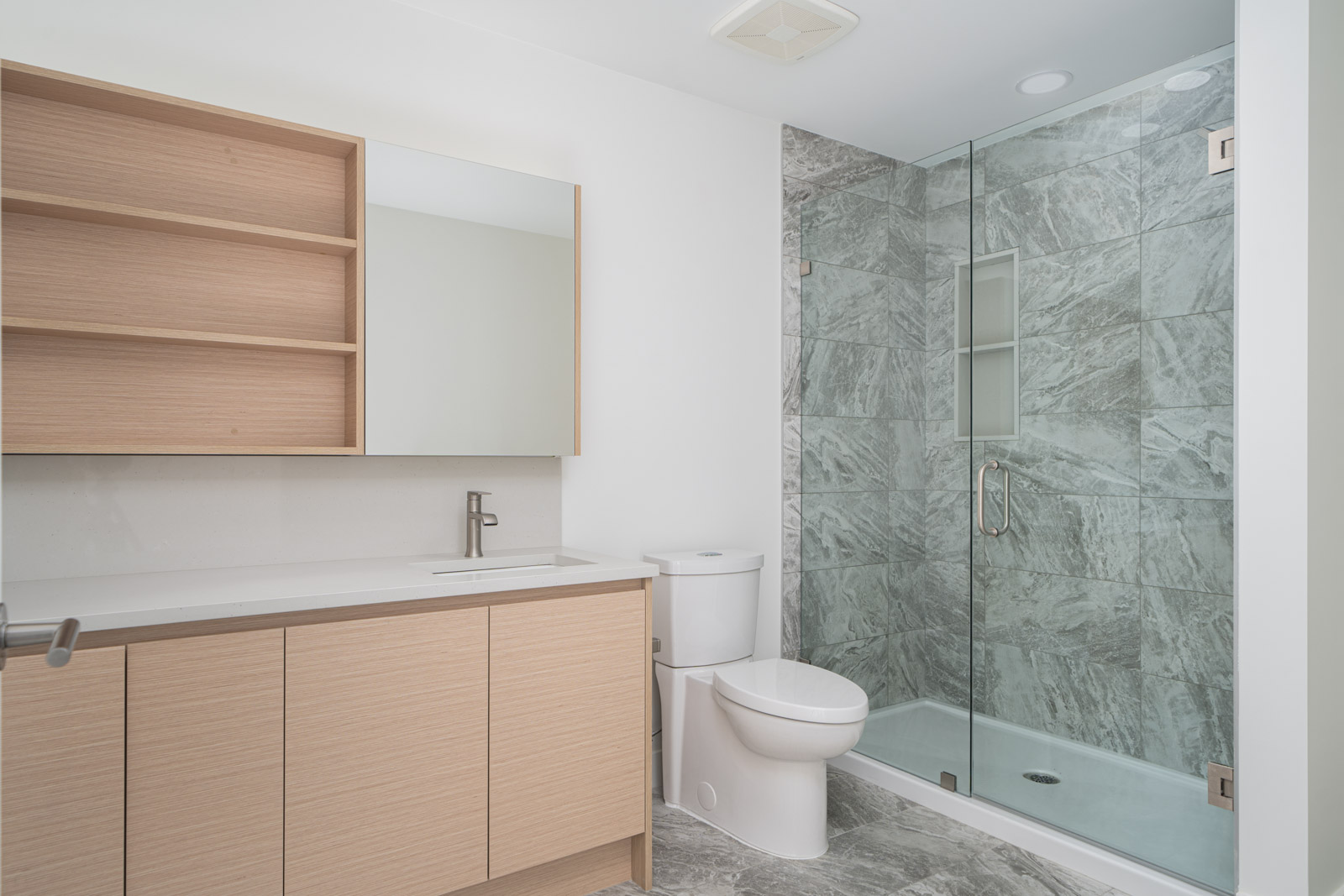 Modern bathroom with light wood cabinetry, a white countertop, a toilet, and a glass-enclosed shower featuring gray marble tile.