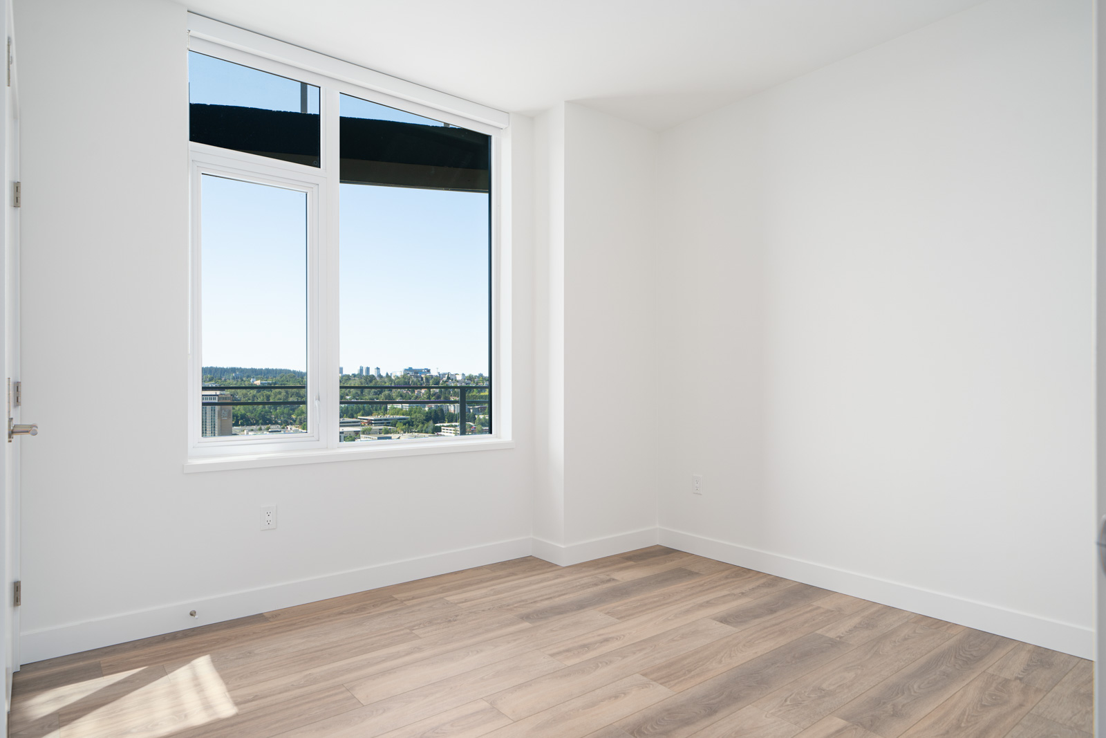 Empty room with white walls, light wood flooring, and a large window showing a view of buildings, greenery, and a clear sky outside.