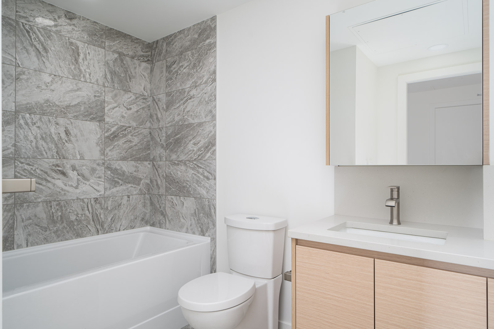Modern bathroom with a white bathtub, gray marble tile wall, a toilet, and a vanity with a sink, mirror, and light wood cabinetry.