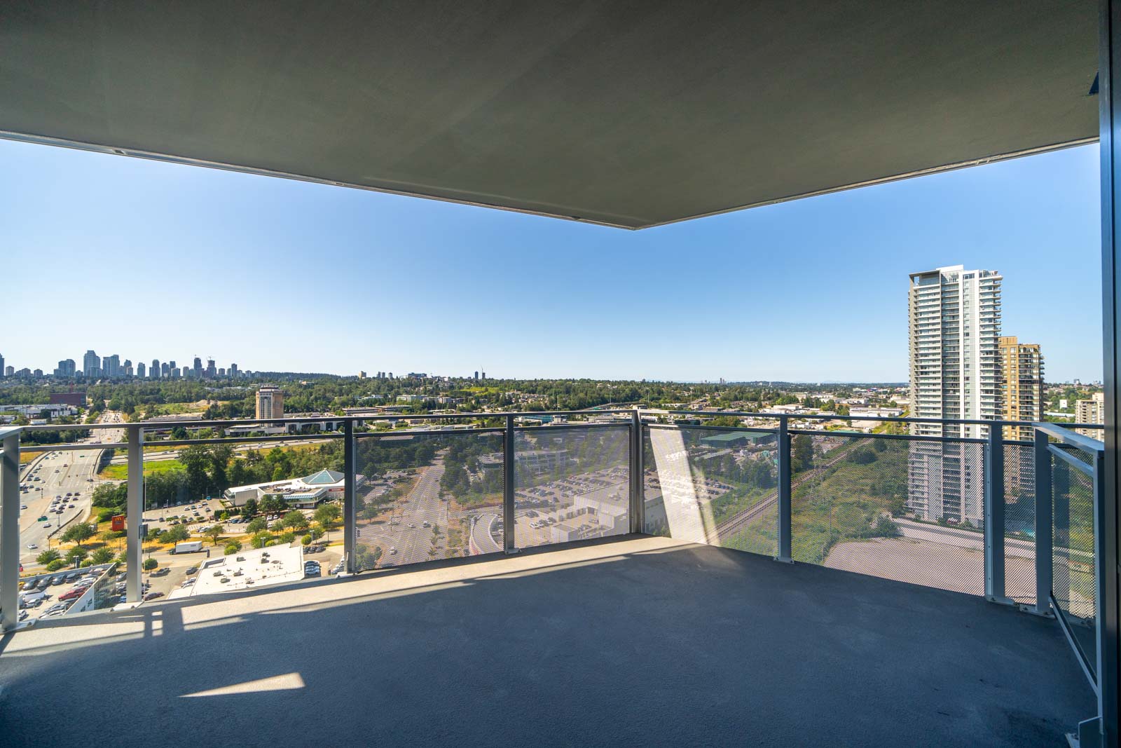 High-rise balcony with glass railing overlooks a cityscape, roads, and green spaces under a clear blue sky.