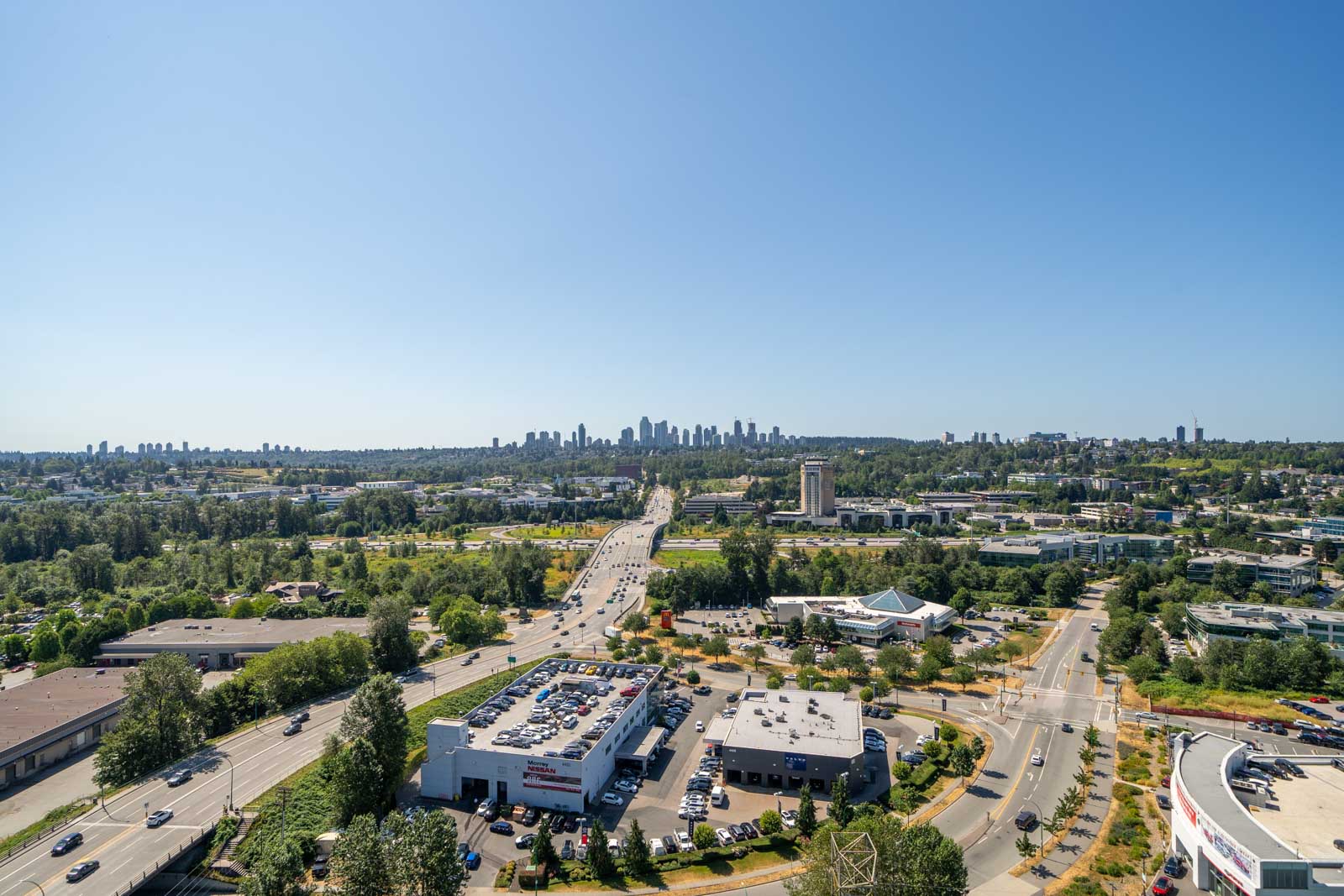A wide view of a suburban area with roads, commercial buildings, and trees, leading towards a distant city skyline under a clear blue sky.