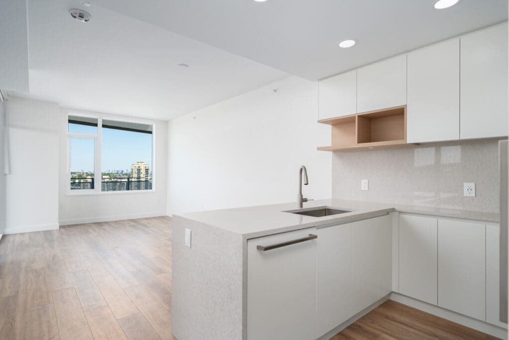 Modern, unfurnished apartment kitchen with white cabinets, a built-in sink, light wood floors, and a large window overlooking city buildings in the background.