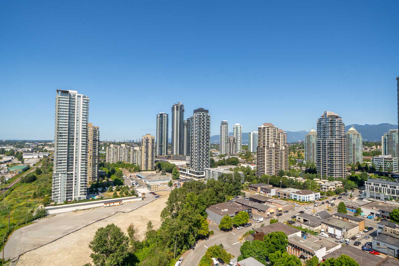 A cityscape featuring numerous high-rise buildings, clear blue sky, distant mountains, and a mix of developed and undeveloped urban areas.
