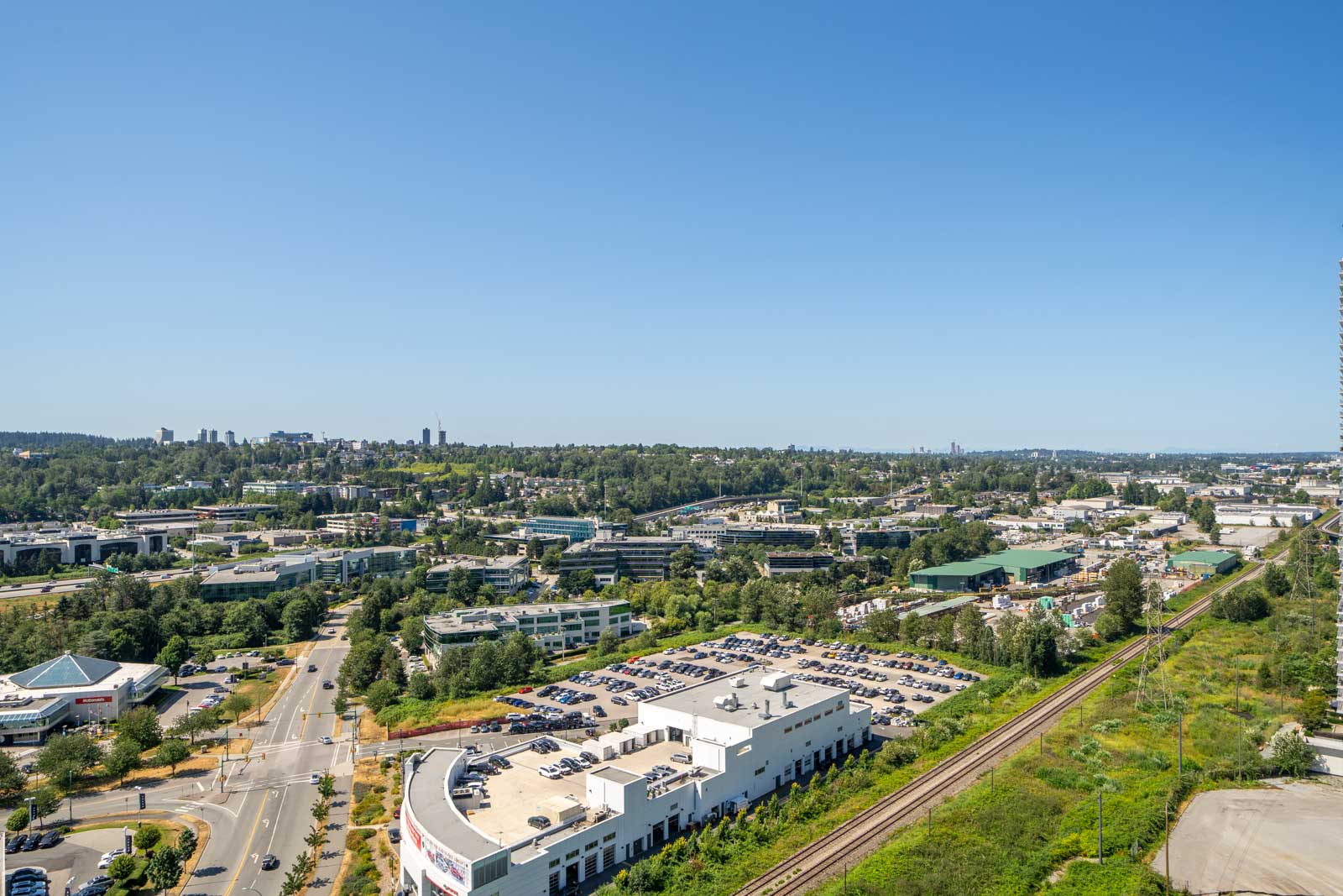 Aerial view of a suburban area with office buildings, parking lots, roads, train tracks, and greenery under a clear blue sky.