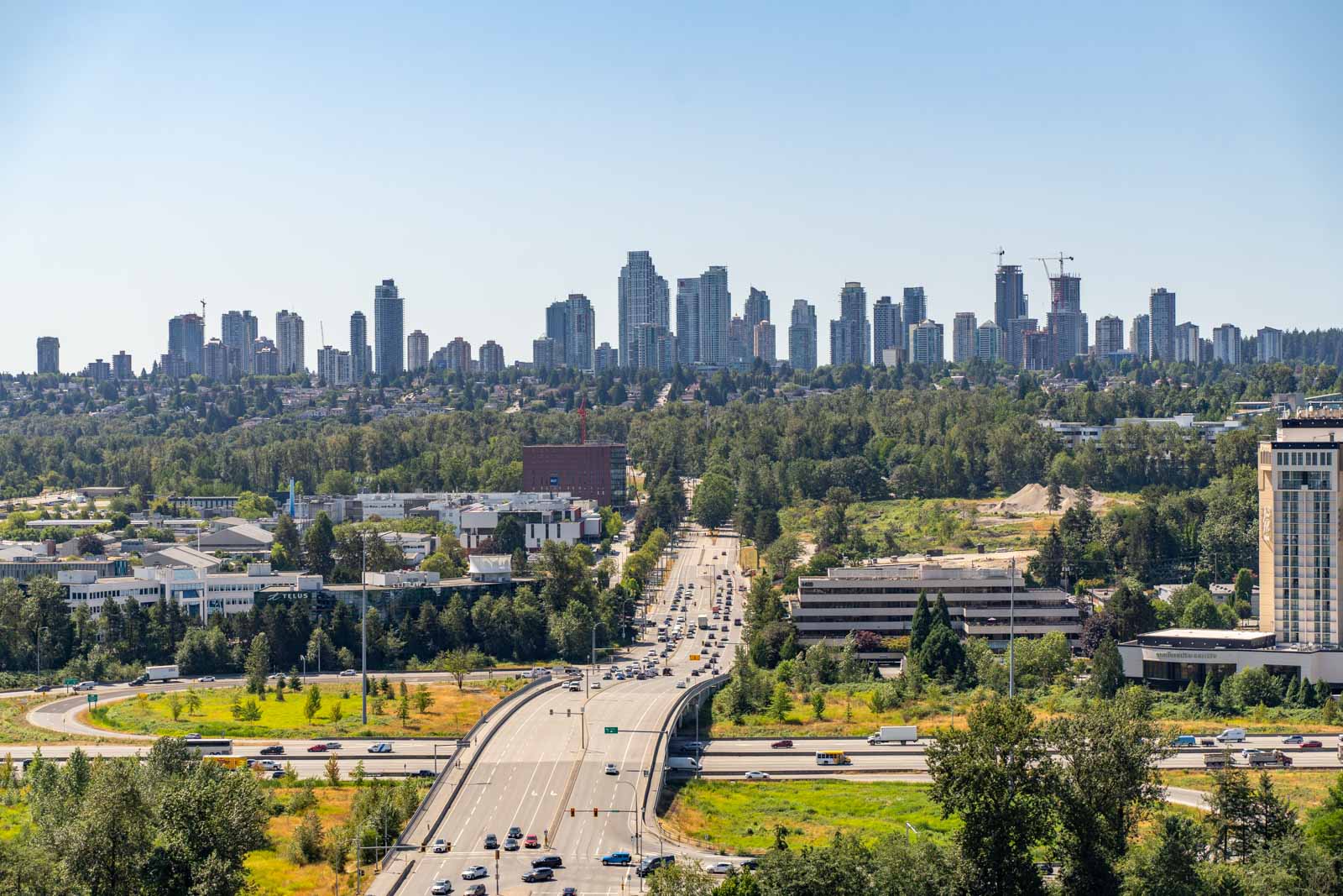 Aerial view of a busy highway surrounded by trees and buildings, with a city skyline of high-rise apartments in the background under a clear blue sky.