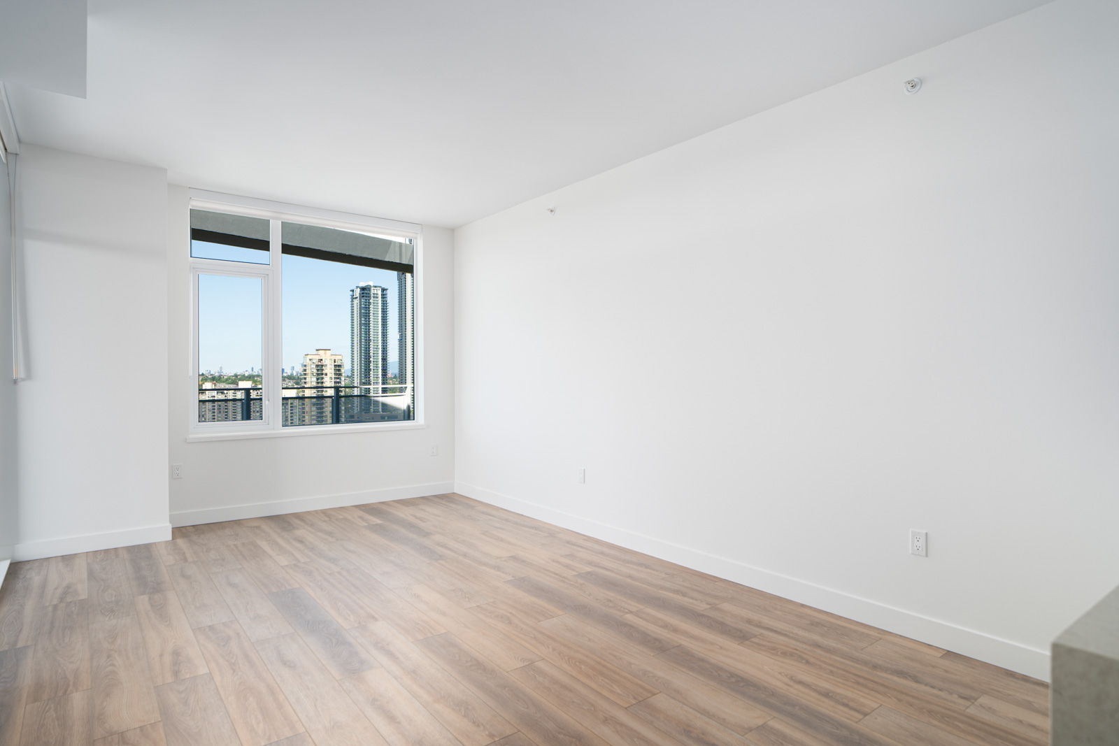 Empty modern room with light wood flooring, white walls, large window, and cityscape view with tall buildings outside.