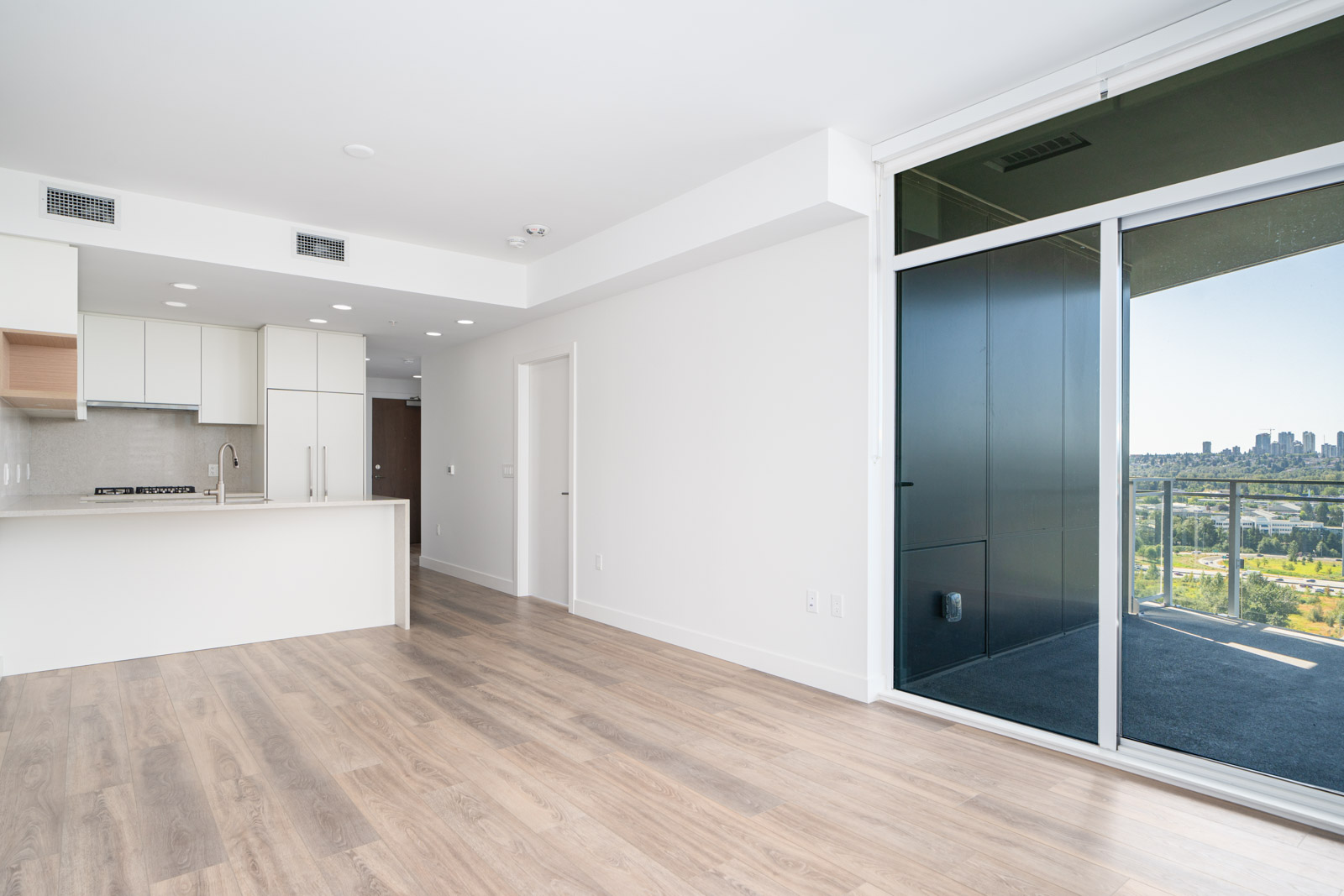 Bright modern apartment interior with light wood flooring, white kitchen, and large glass doors opening to a balcony with a city view.