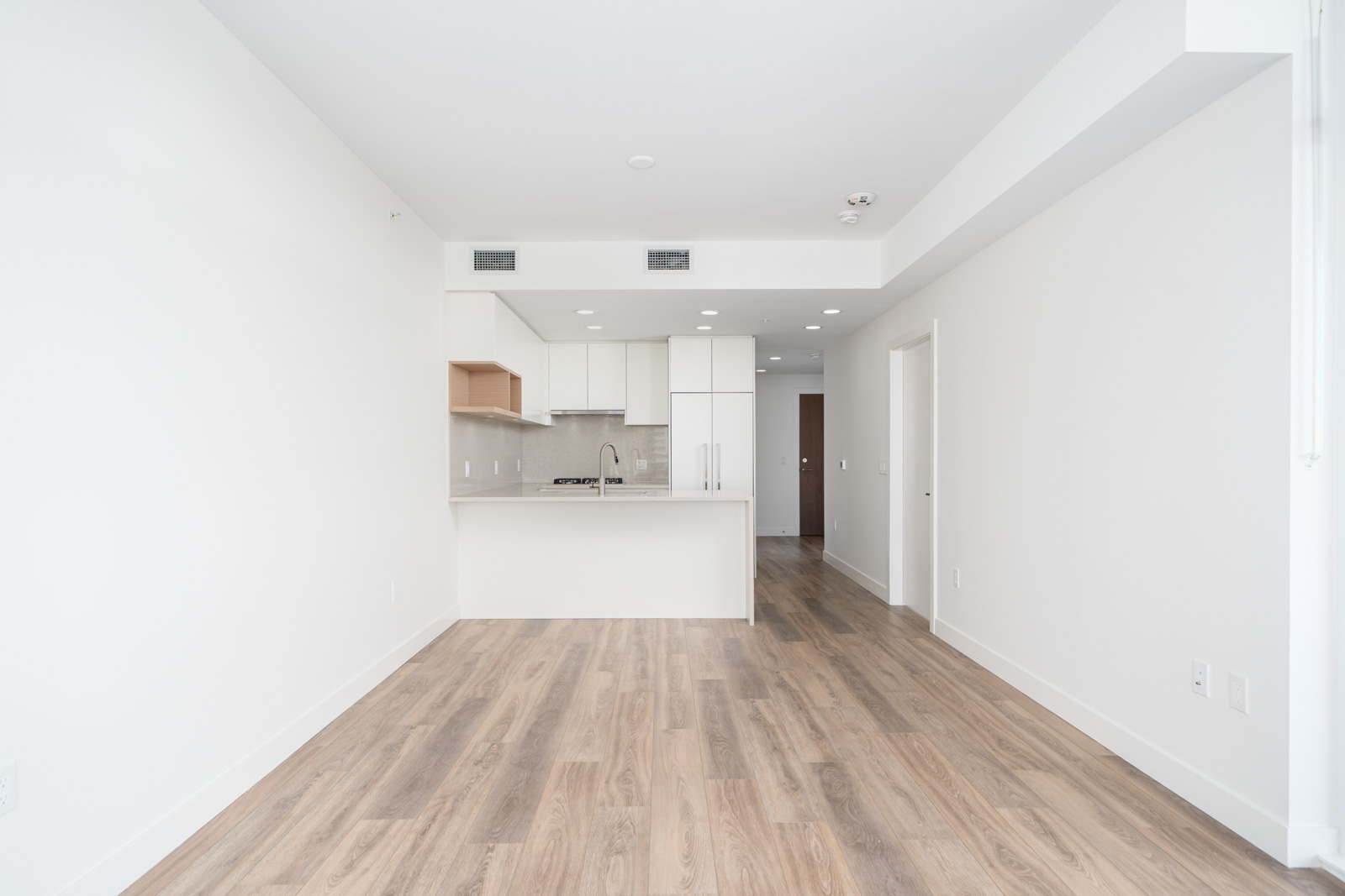 Modern, unfurnished apartment with white walls, light wood flooring, and an open-plan kitchen featuring white cabinets and a center island.