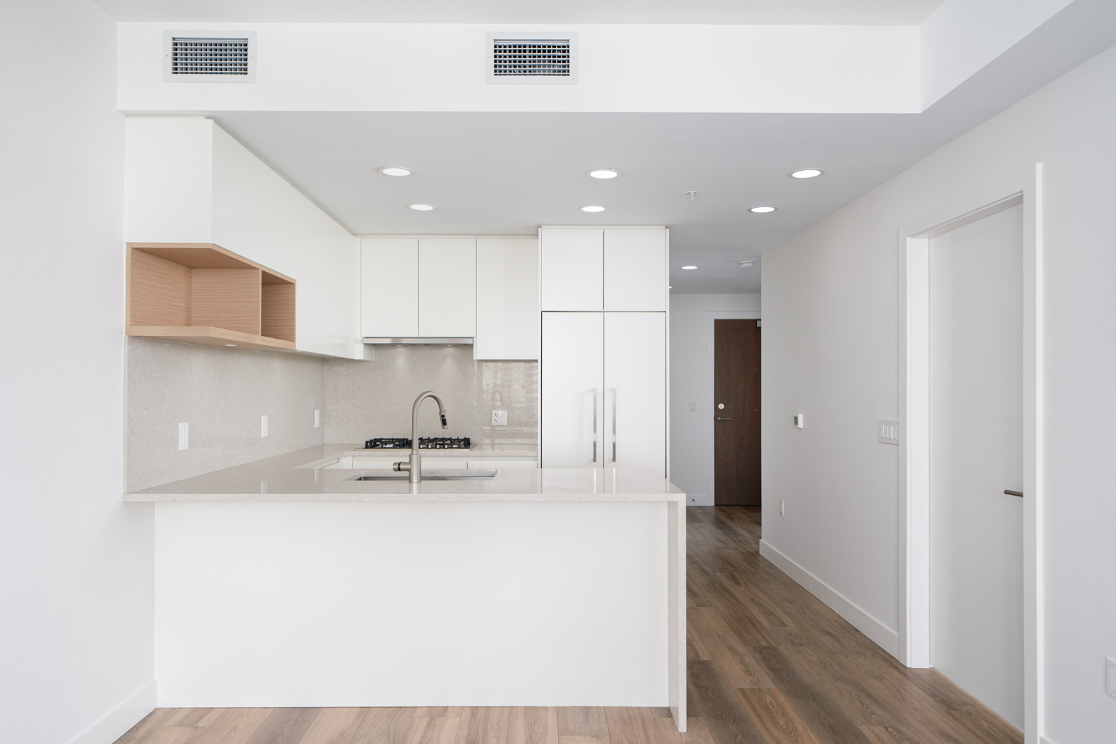 Modern, minimalistic kitchen with white cabinets, a built-in stove, light wood flooring, and an island sink; open layout leads to entry door in the background.