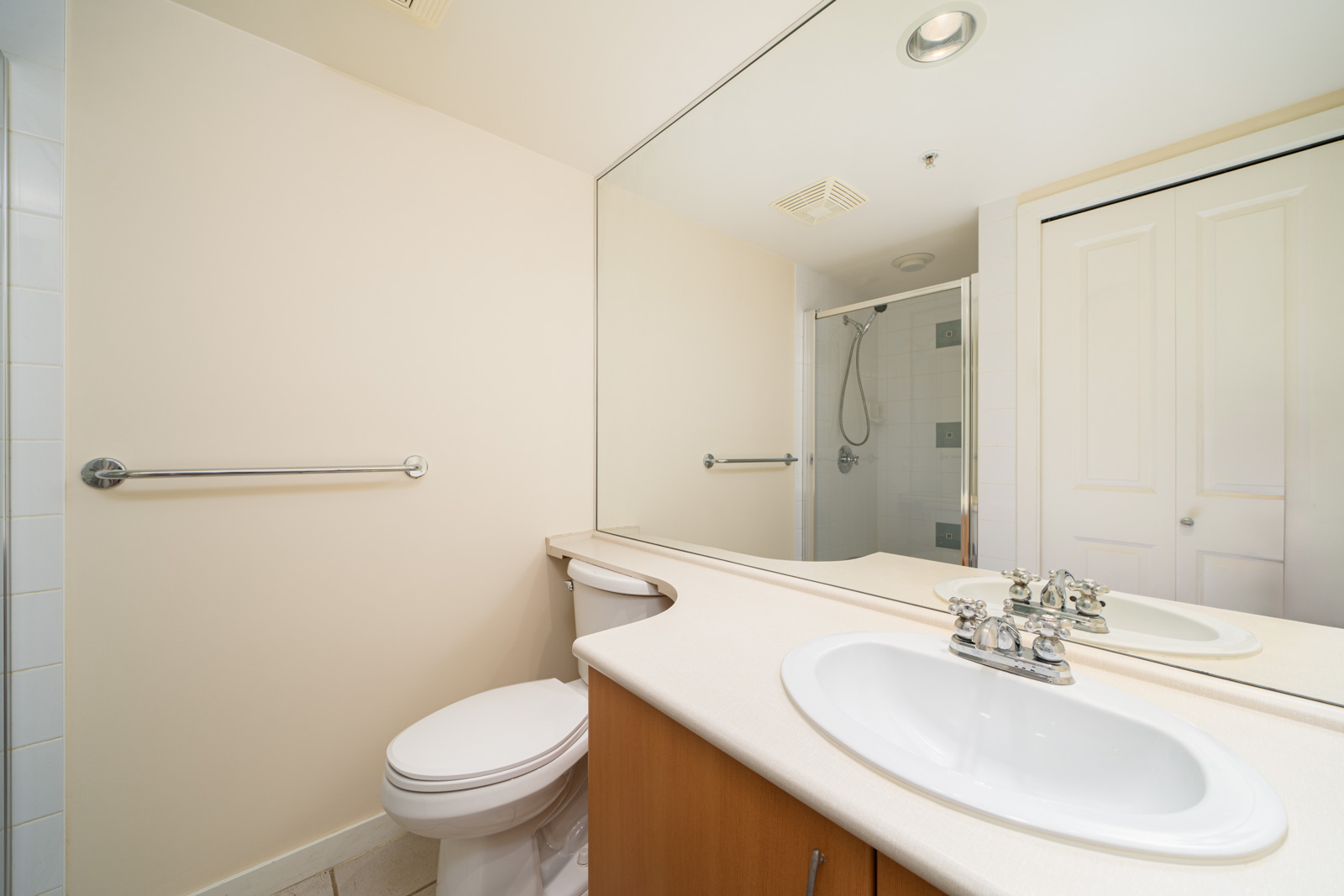 A clean bathroom with a white sink, countertop, toilet, large mirror, towel bar, and a shower with a glass door in the background.