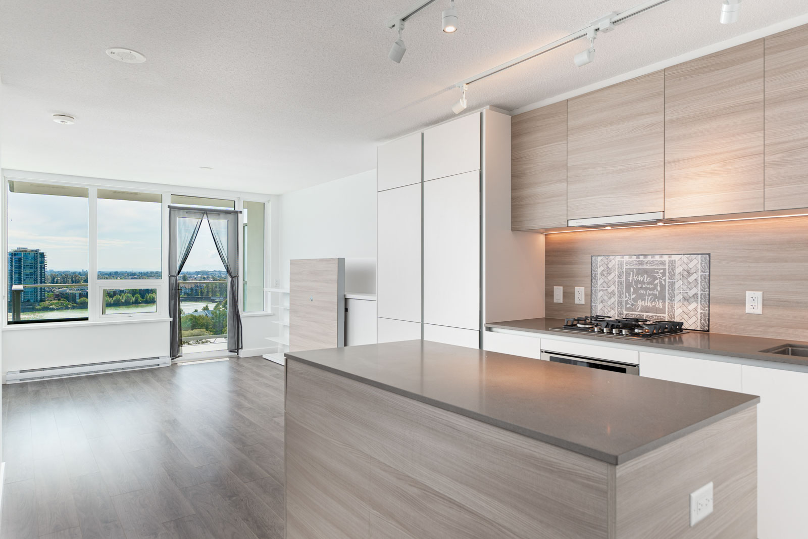 Modern open-concept kitchen with light wood cabinets, gray countertops, built-in appliances, and large windows overlooking a cityscape.