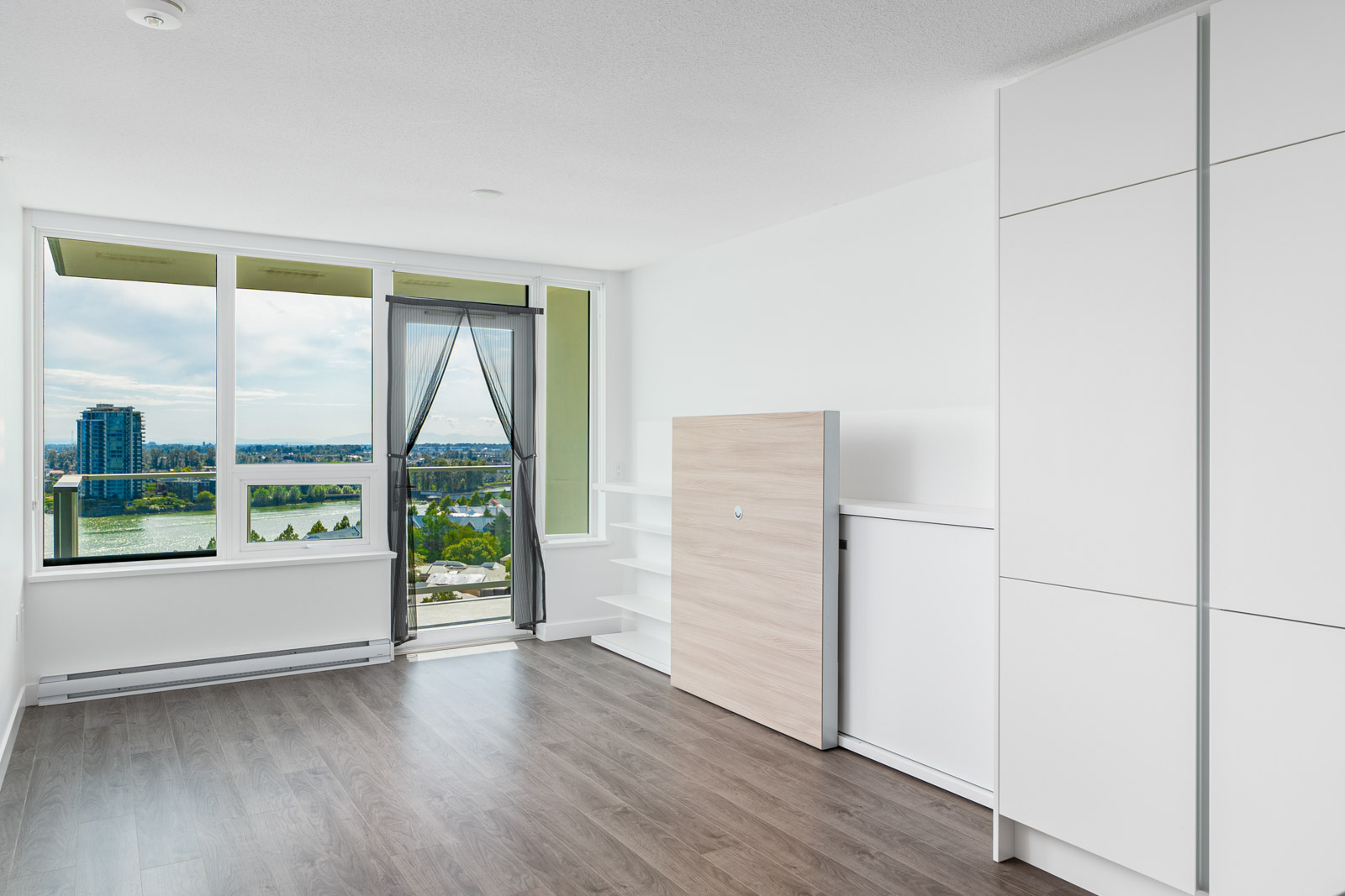Bright, unfurnished modern apartment room with large windows, built-in shelving, white cabinets, wood flooring, and balcony view of a river and buildings.