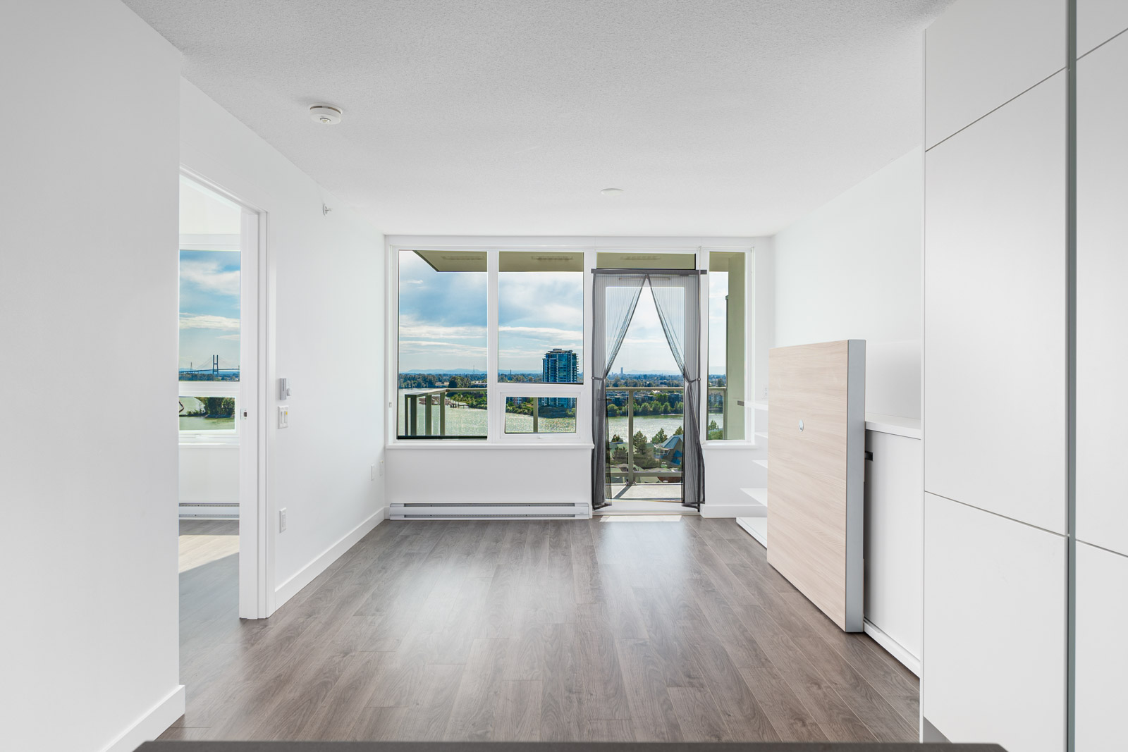Unfurnished modern apartment with large windows, balcony door, wood flooring, and a partial city and water view in the background.