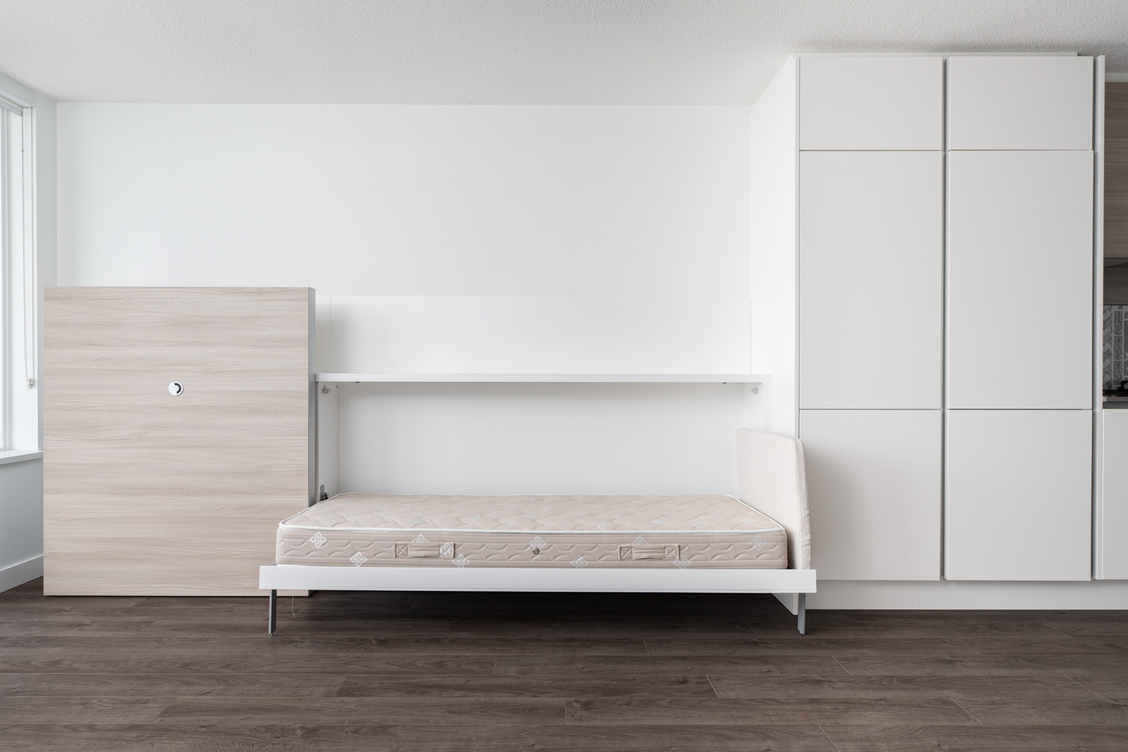 A modern, minimalist room features a folded-down Murphy bed with a bare mattress, white shelving, and built-in cabinets against a white wall and dark wood flooring.