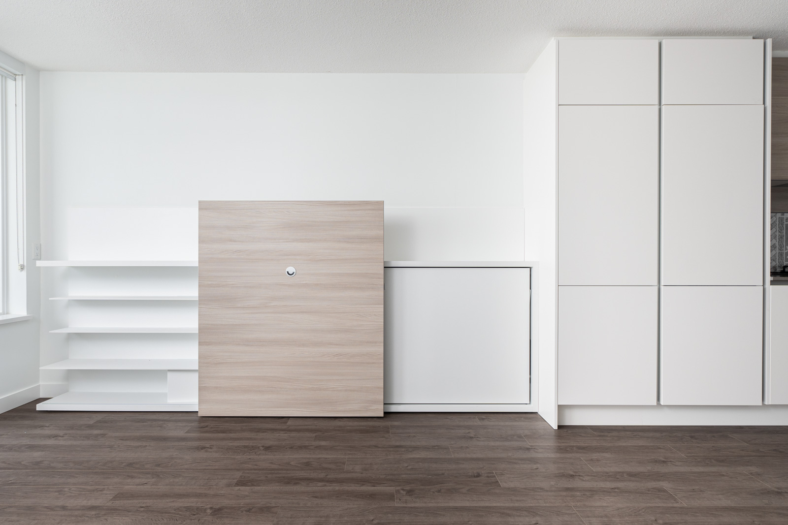 Minimalist room with white built-in shelves and cabinets, light wood flooring, and a large light wood panel in front of the shelving.