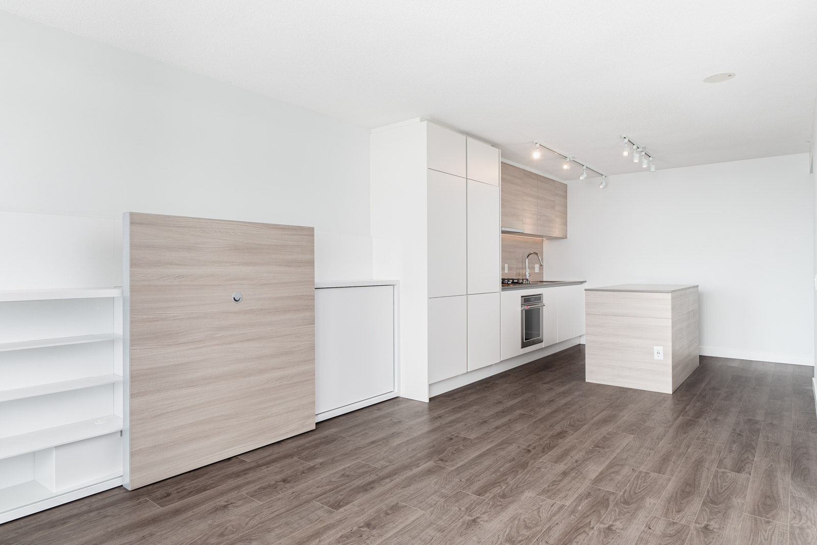 Minimalist modern kitchen with light wood cabinets, white countertops, an island, built-in oven, and dark wood flooring in an open, unfurnished space.