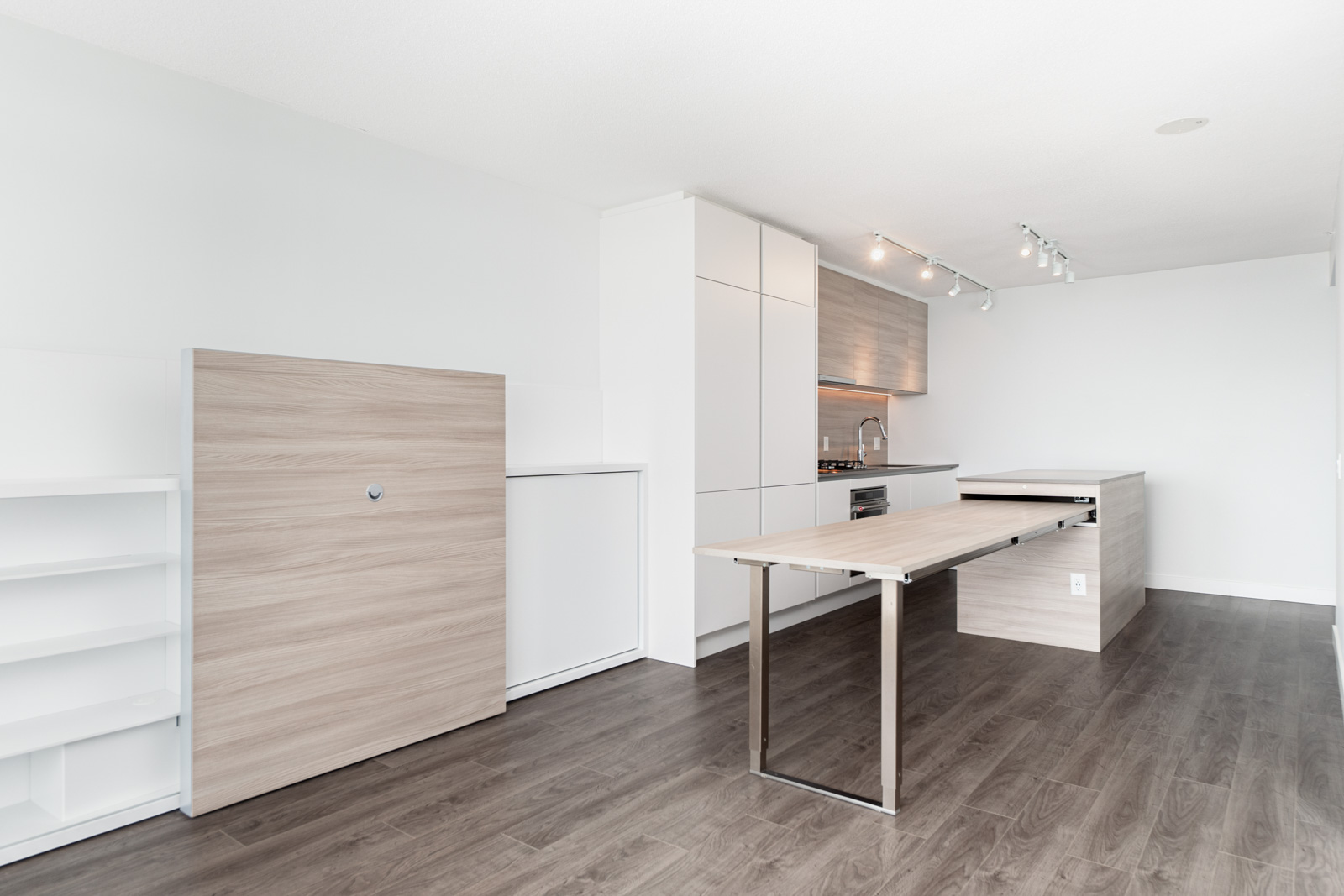 Minimalist kitchen with white cabinets, wood accents, pull-out table, open shelving, and dark wood flooring under track lighting.