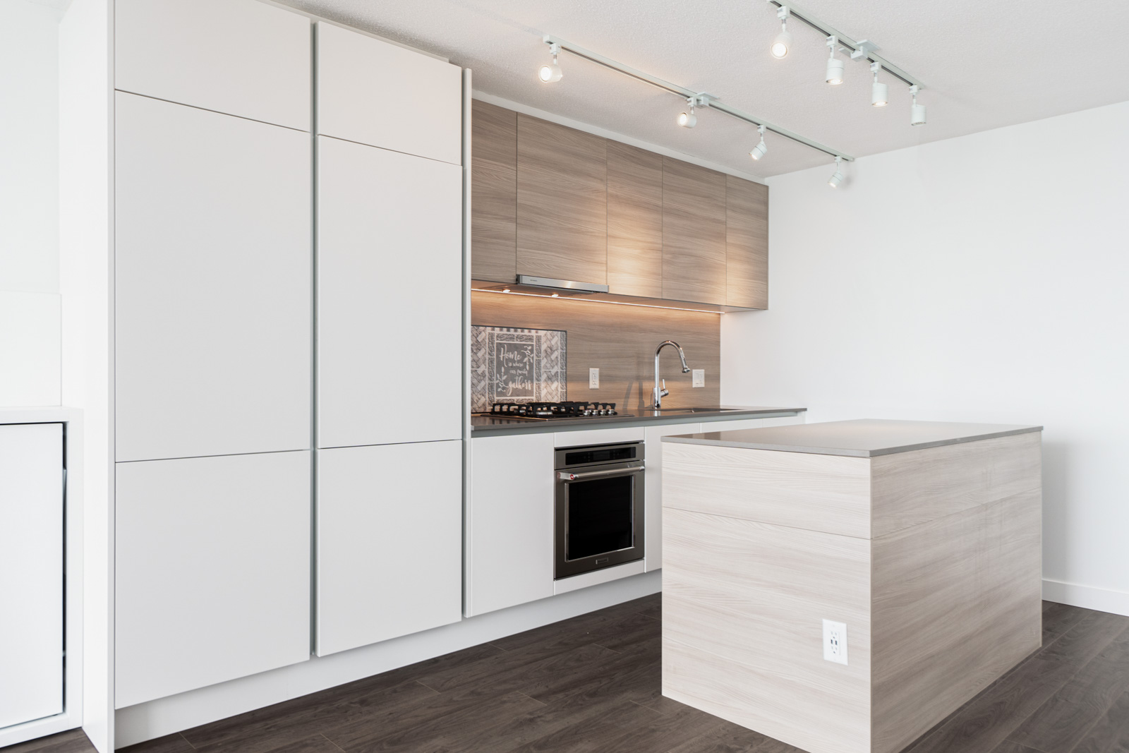 Minimalist kitchen with light wood cabinets, a small island, built-in oven, stovetop, sink, and track lighting; features white and wood finishes with dark wood flooring.
