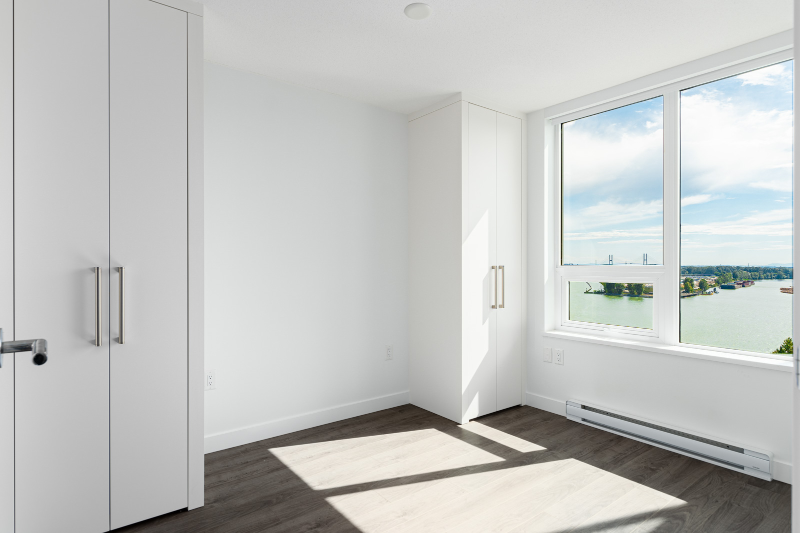 Bright, empty room with white walls, built-in closets, and large window overlooking a water view and blue sky.