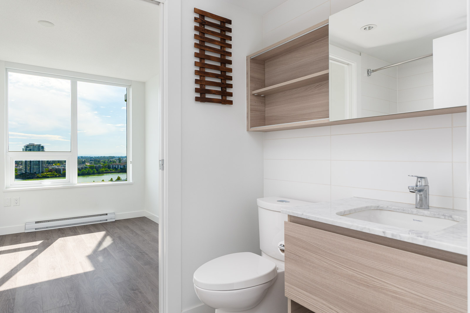 A modern bathroom with light wood cabinets, a white toilet, and a marble countertop, next to a bright room with large windows and a city view.