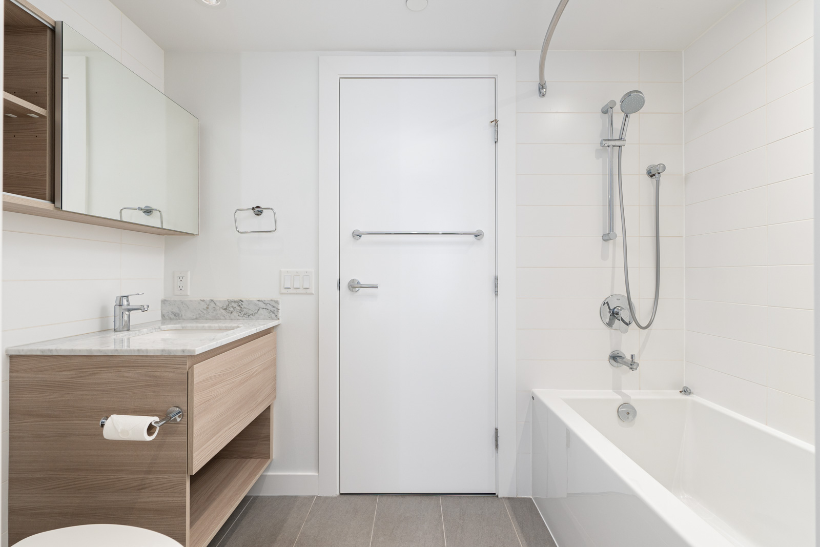 Modern bathroom with a white bathtub, handheld shower, wooden vanity with sink, wall mirror, and towel rail on a white door. Minimalist design with neutral colors.