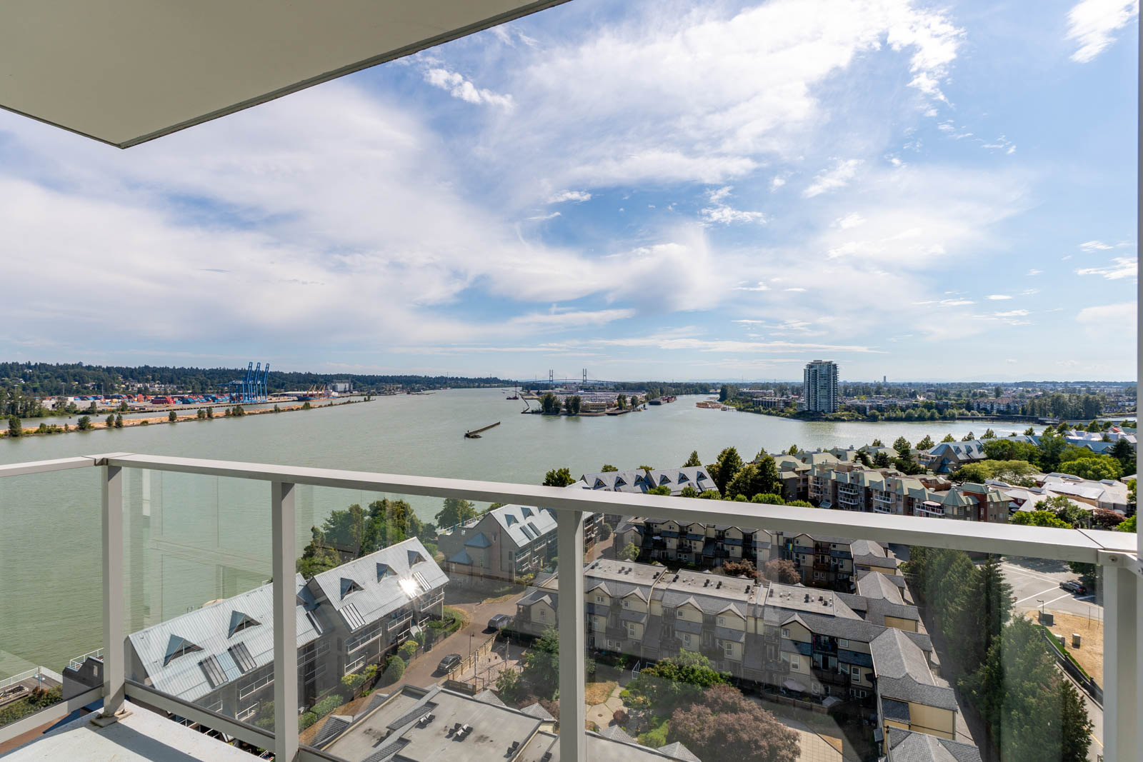 View from a high-rise balcony overlooking a river, residential buildings, and a distant bridge under a partly cloudy sky.
