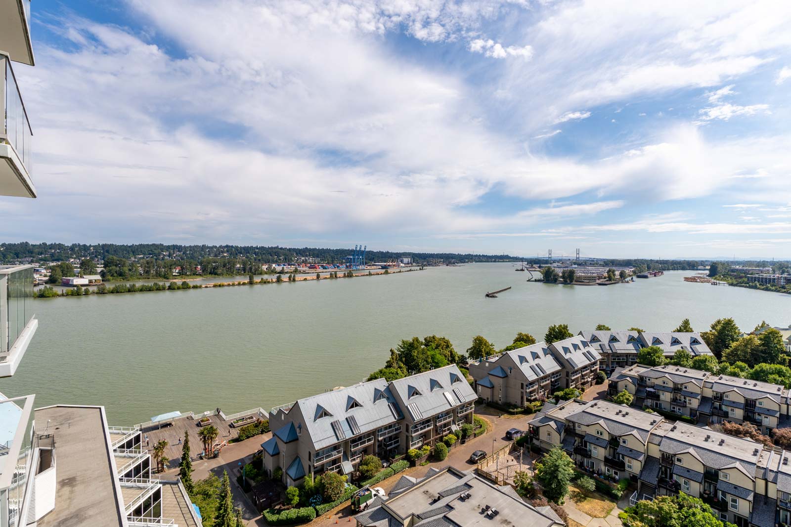 Aerial view of a river with a barge, rows of townhouses in the foreground, and trees along the shoreline under a partly cloudy sky.