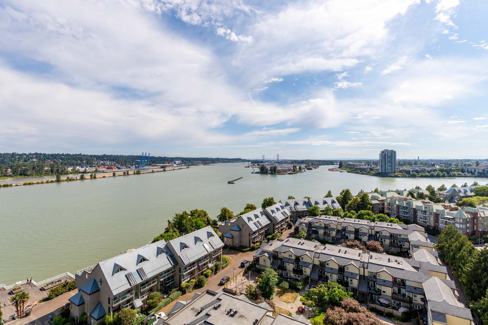 Aerial view of a riverside residential area with apartment buildings, trees, and a wide river under a partly cloudy sky.