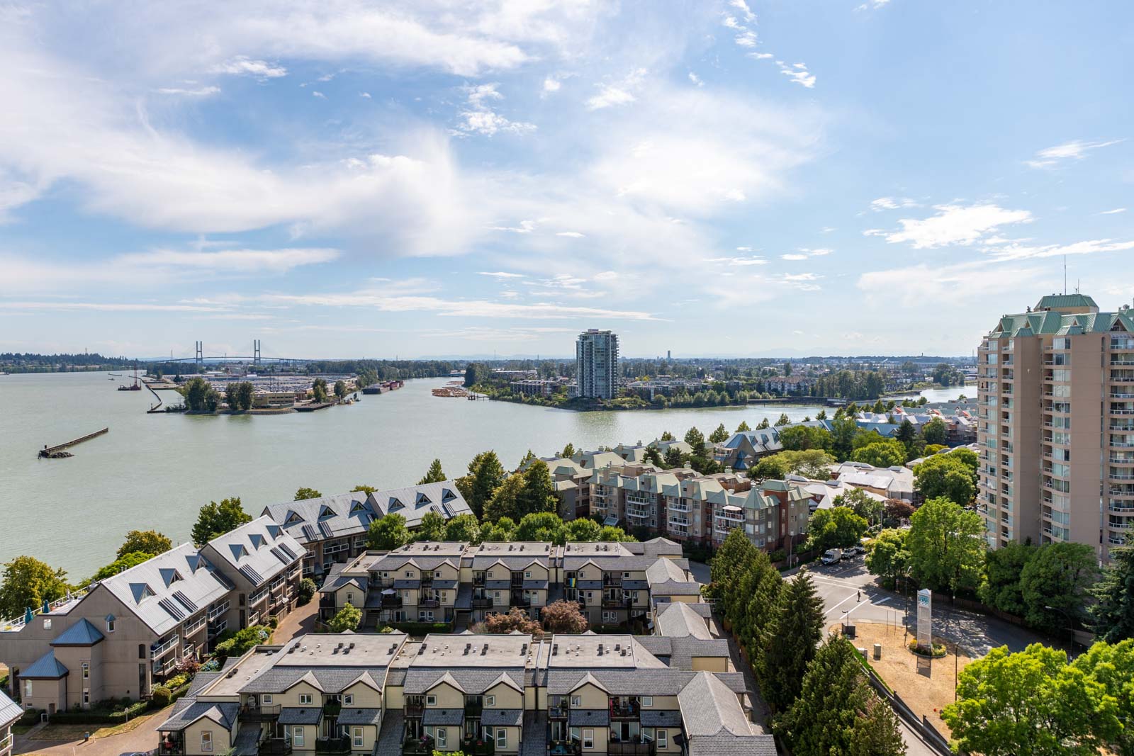 Aerial view of residential buildings and townhouses near a river, with a bridge, boats, and high-rise apartments in the background under a partly cloudy sky.