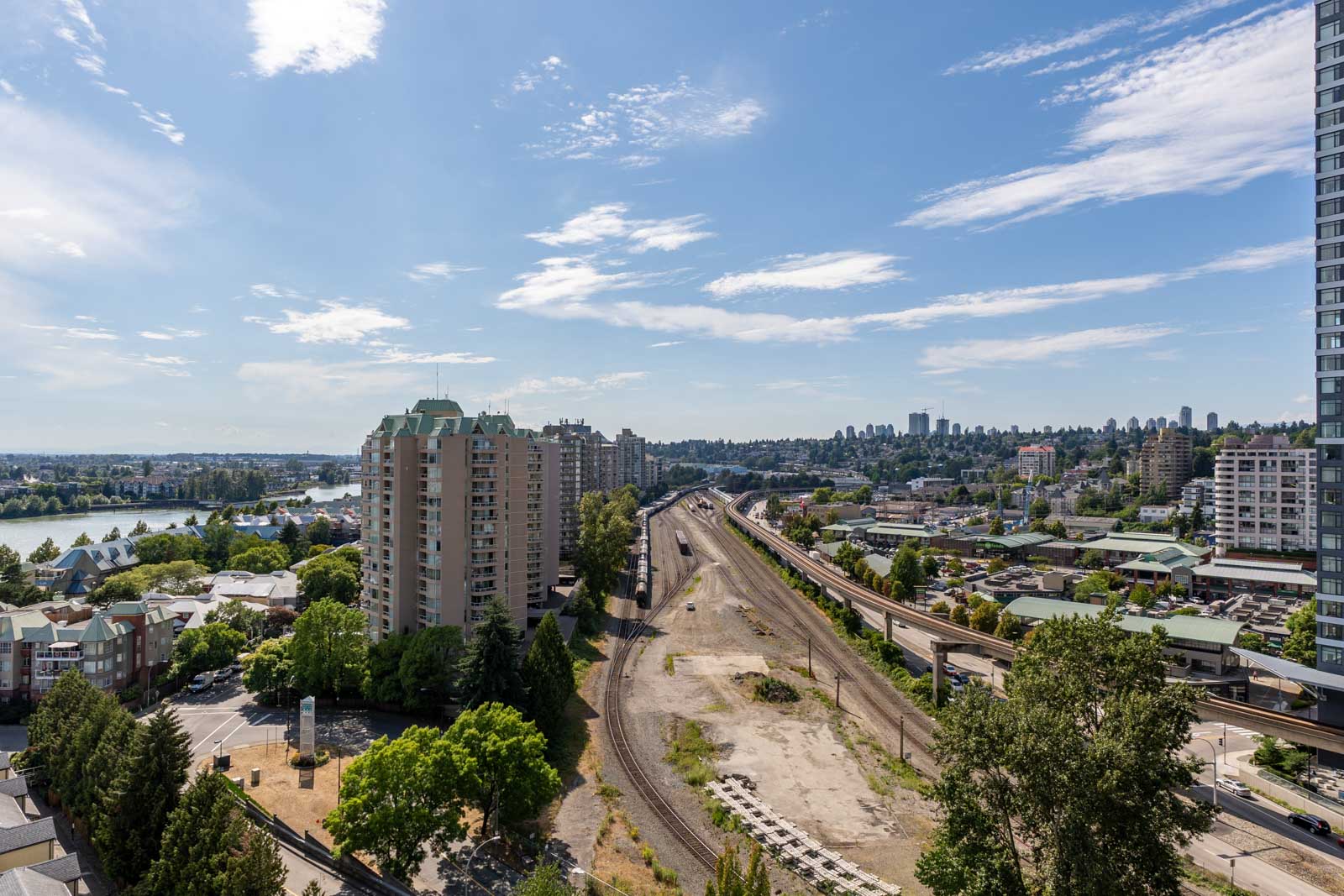 A cityscape showing residential buildings, railway tracks, and a mix of greenery under a blue sky with scattered clouds.