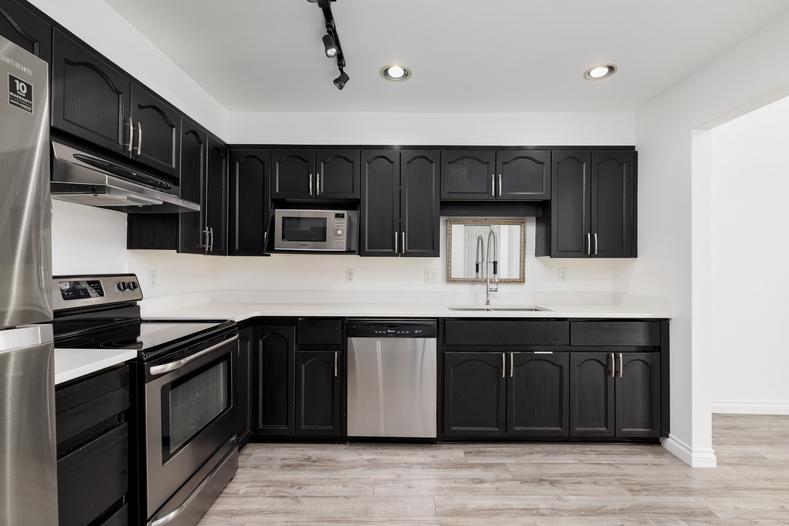 Modern kitchen with black cabinets, stainless steel appliances, white countertops, light wood flooring, and track lighting on the ceiling.
