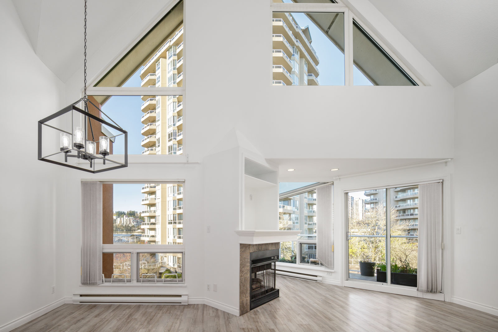 A modern, unfurnished living room with large windows, high ceilings, a fireplace, and pale wood flooring, overlooking tall apartment buildings outside.