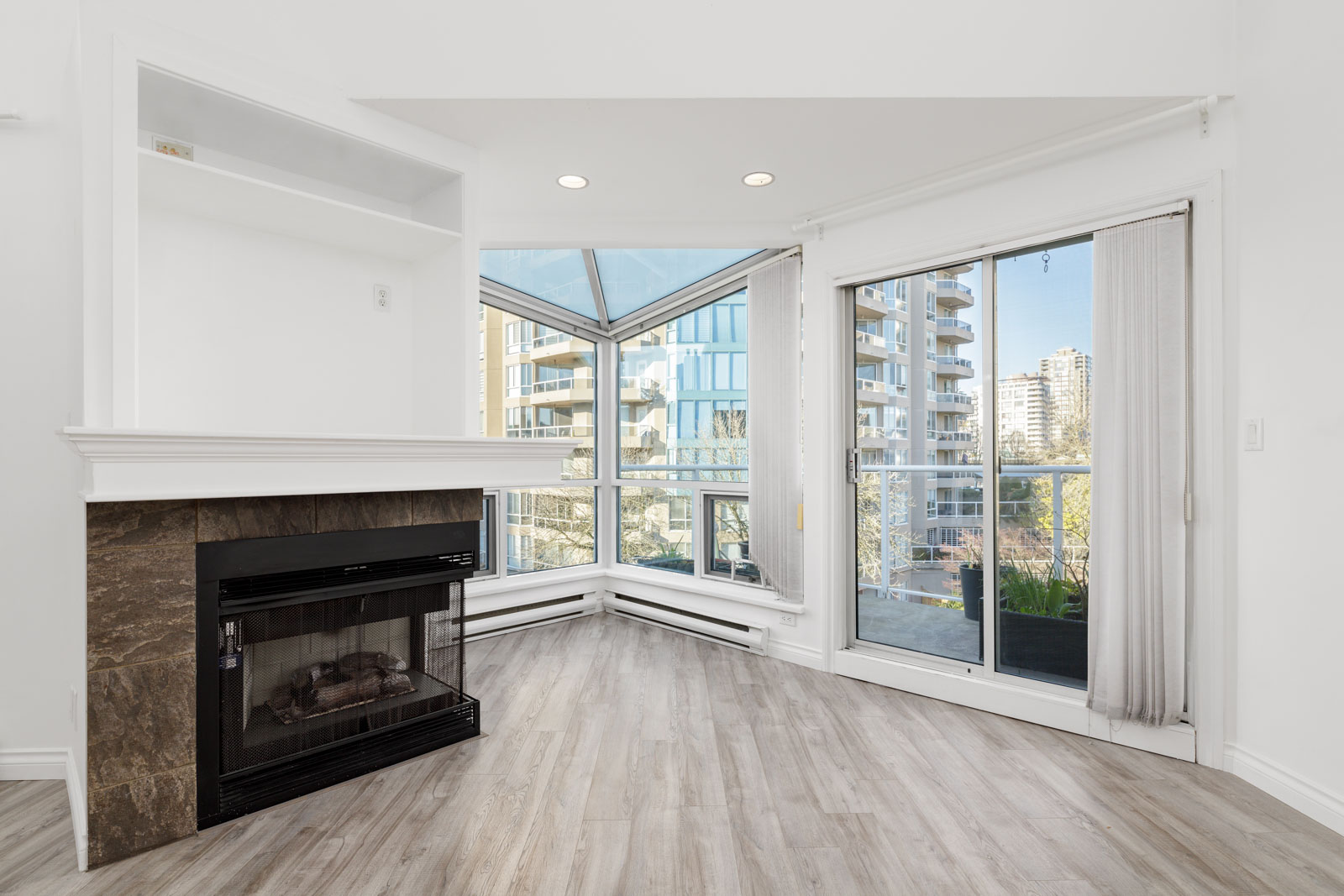Bright modern corner room with large windows, a sliding glass door to a balcony, a fireplace with a stone base, and light wood flooring.