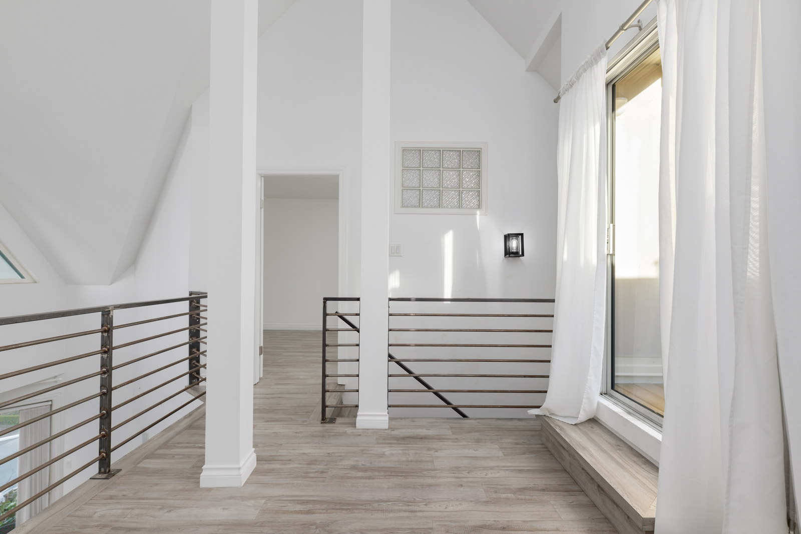 A modern loft landing with light wood flooring, white walls, metal railings, and a large window with sheer curtains letting in natural light.