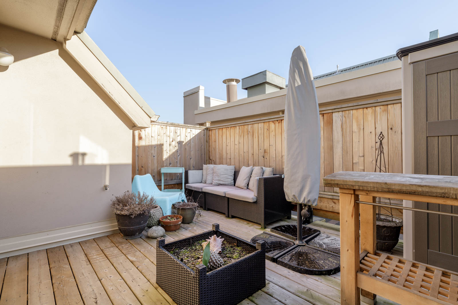 A wooden rooftop patio features outdoor seating with cushions, a small table, potted plants, an umbrella, and a wooden privacy fence under a clear sky.