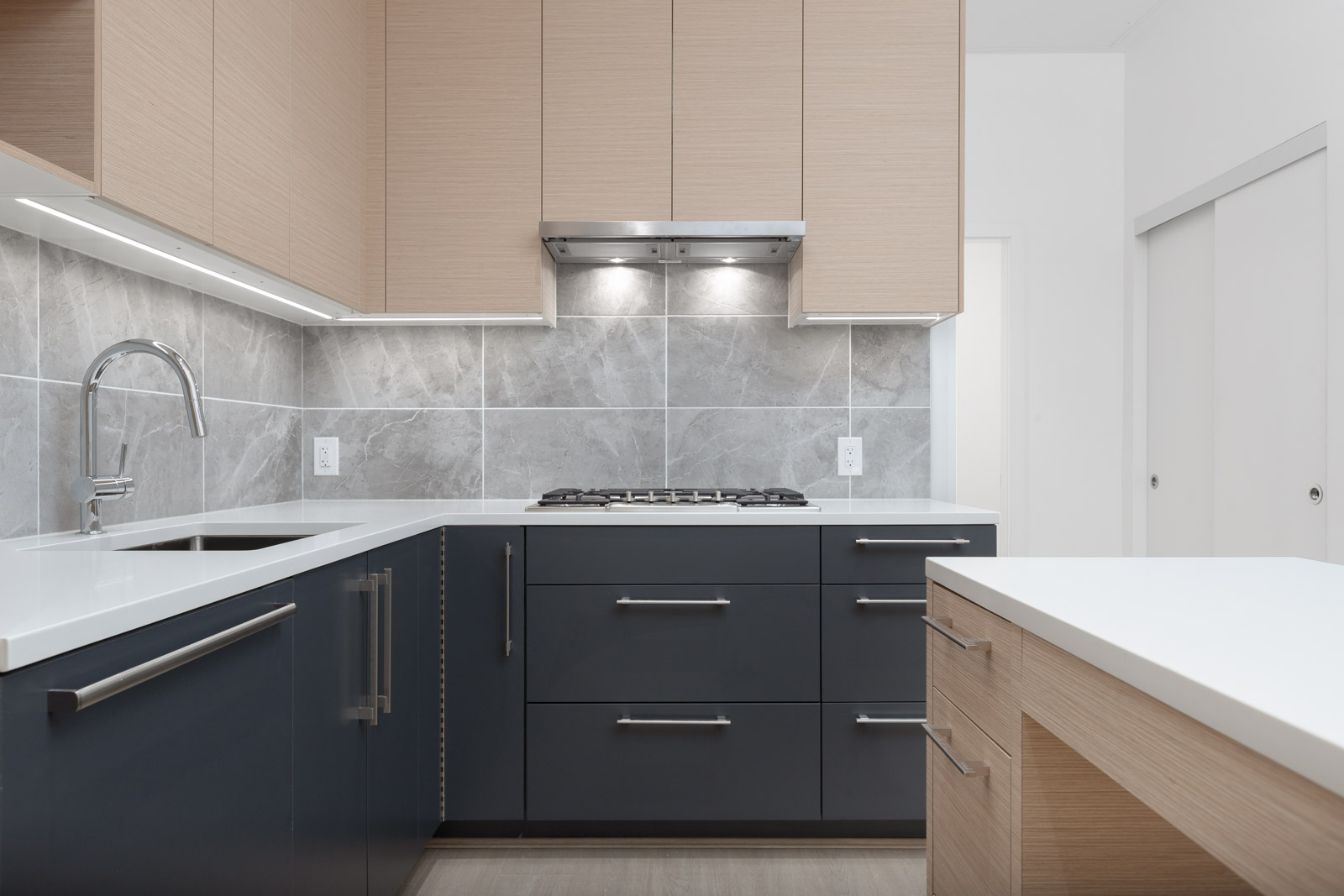 Modern kitchen with light wood upper cabinets, dark lower cabinets, stainless steel handles, a gas cooktop, under-cabinet lighting, and gray tile backsplash.
