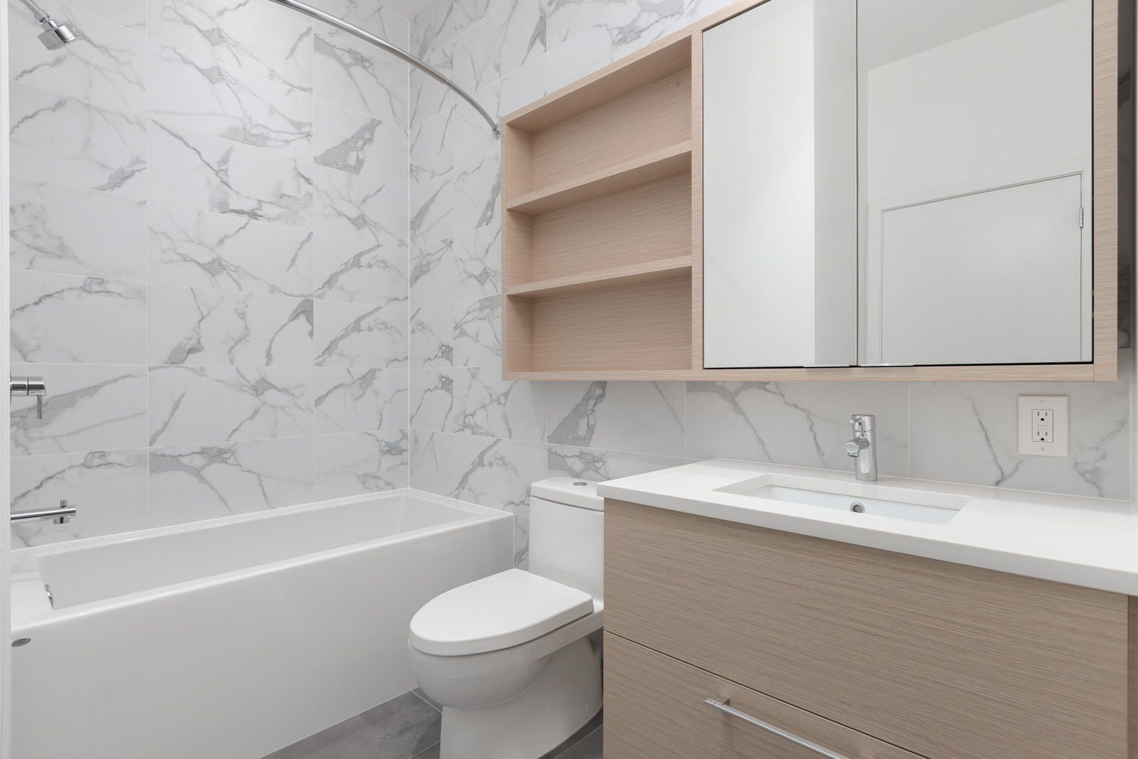 Modern bathroom with a white bathtub, toilet, sink, wood cabinets, open shelves, and mirrored medicine cabinet, featuring marble-patterned tile walls.