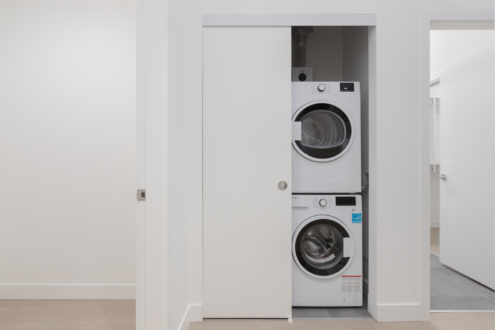 A white stacked washer and dryer set is placed inside a small closet with a sliding door in a modern, minimalistic room.