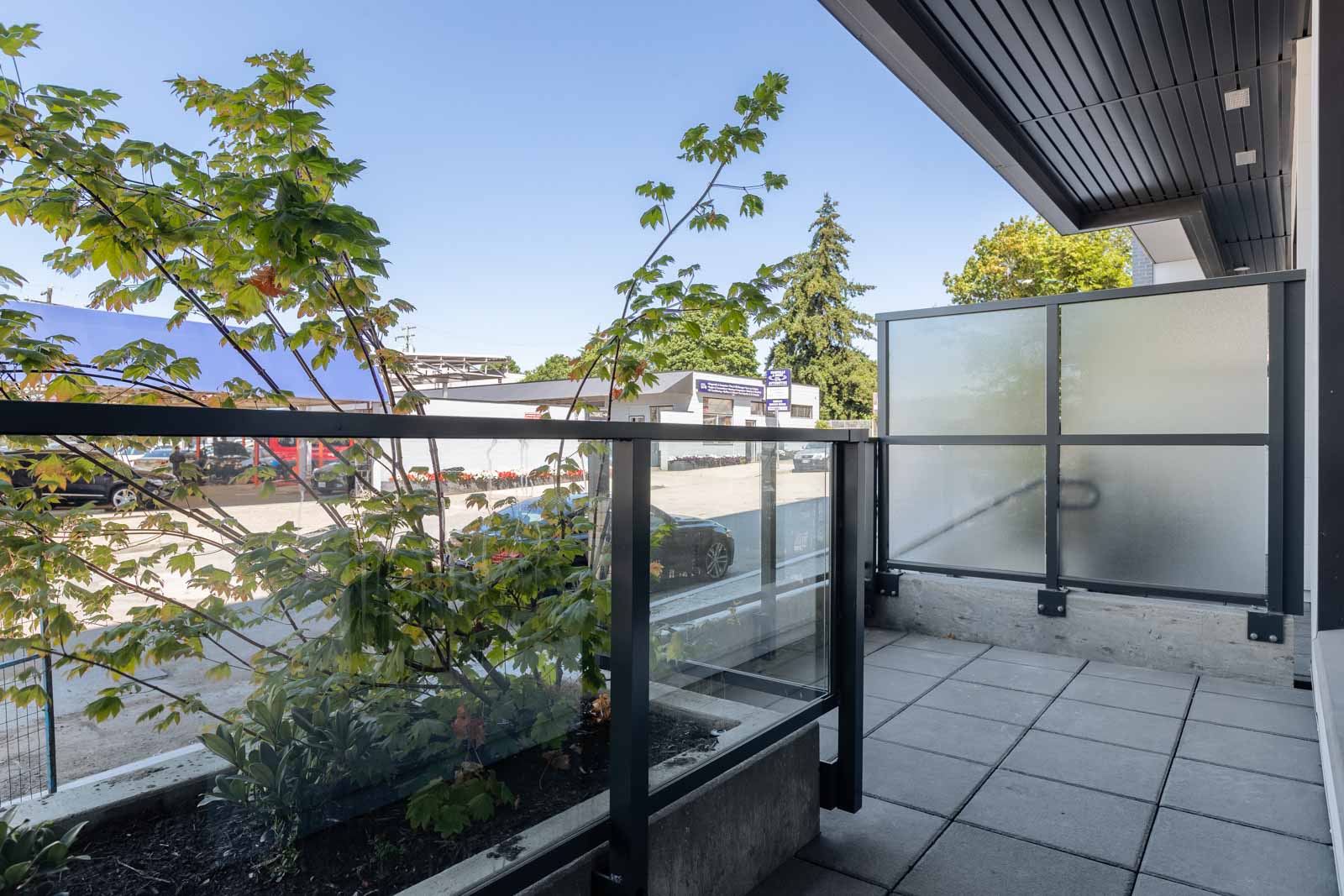 A small balcony with glass railing, frosted privacy panel, potted plant, and tiled floor overlooking a parking lot and commercial buildings.