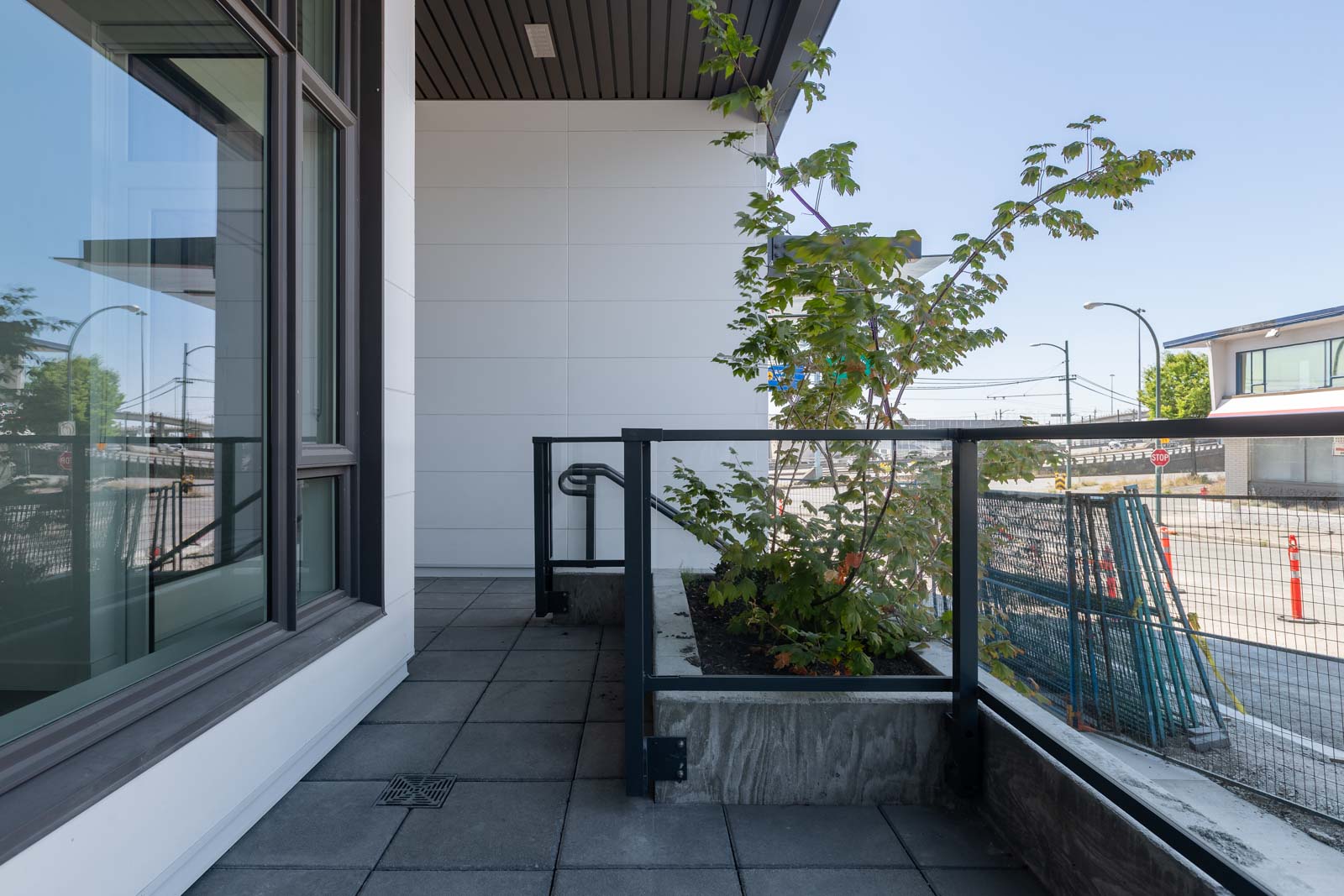A modern balcony with tiled flooring, glass railing, and a small planter with a leafy green plant; urban street and buildings visible in the background.