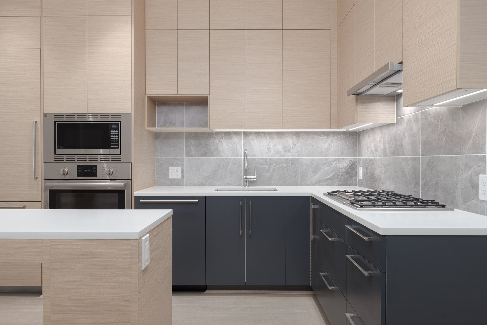 Modern kitchen with light wood cabinets, gray lower cabinets, built-in oven and microwave, gas cooktop, white countertops, and gray tile backsplash.