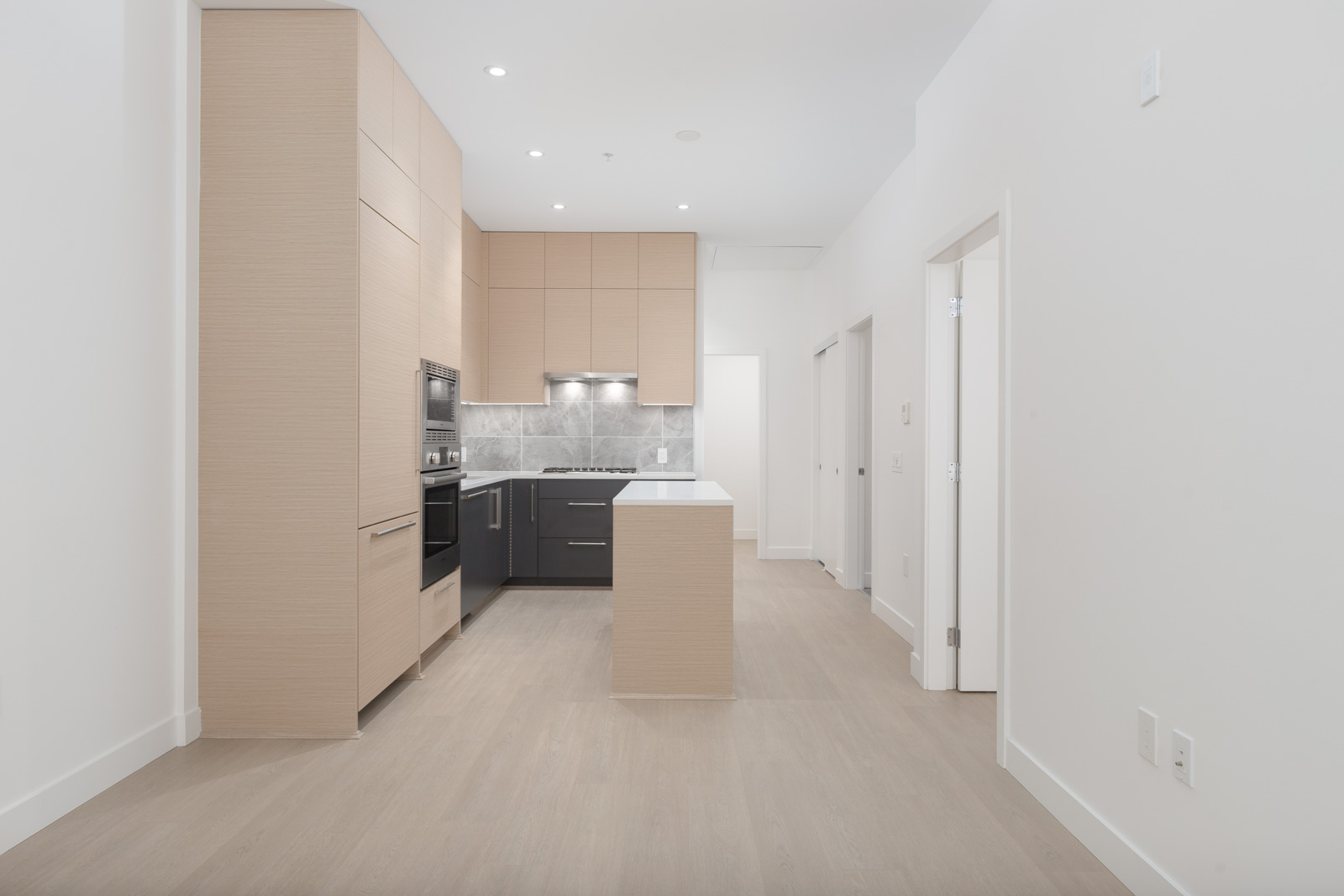 Modern kitchen with light wood cabinets, grey backsplash, black appliances, and a small central island, viewed from an open, unfurnished space with light wood flooring.