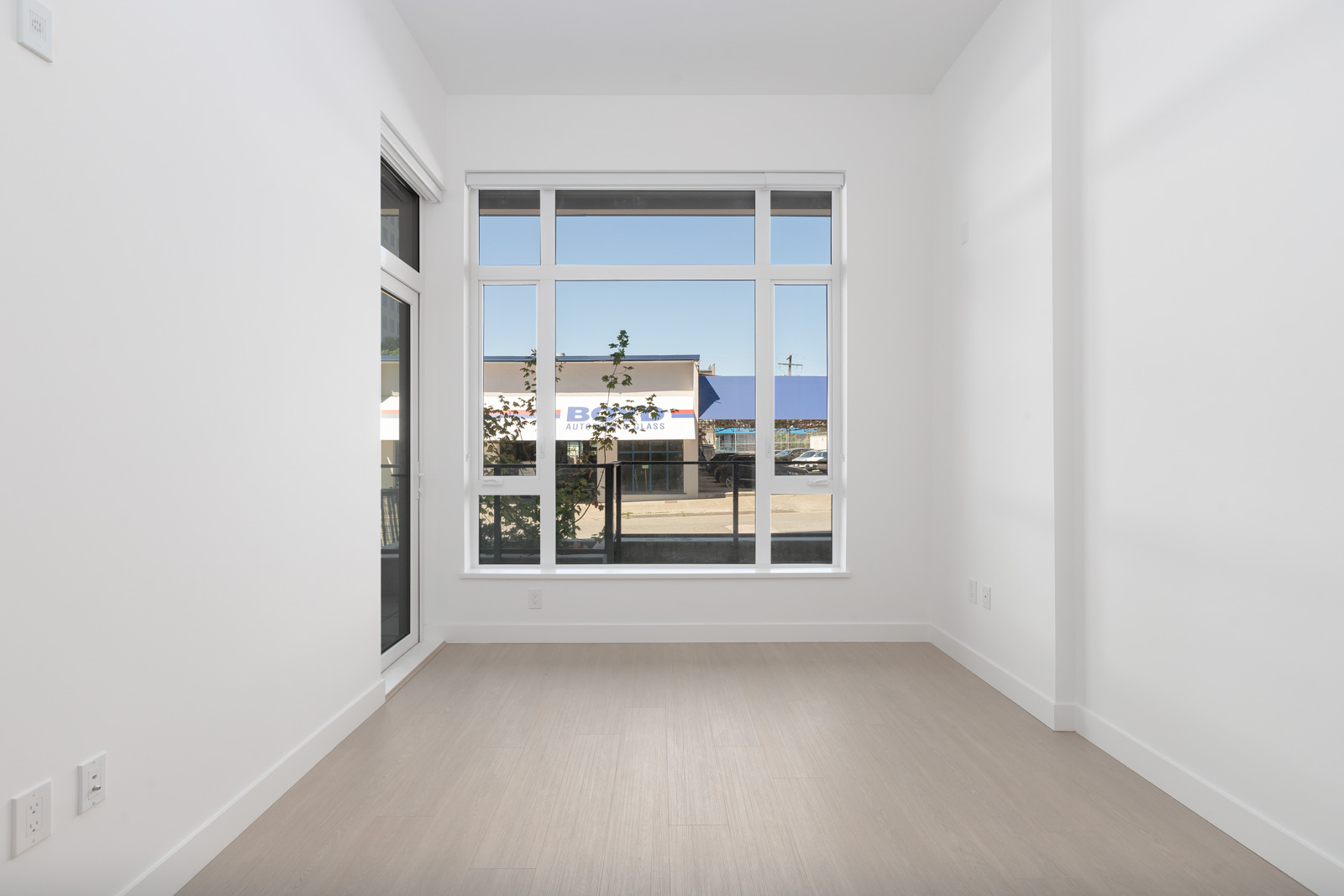 Empty, unfurnished room with large windows, white walls, light wood flooring, and a view of a building and street outside.