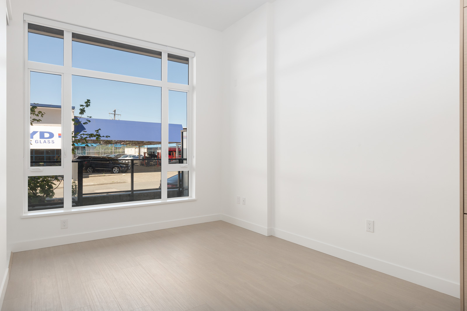 Empty room with light-colored wood flooring, white walls, and a large window overlooking a street with parked cars and a building outside.