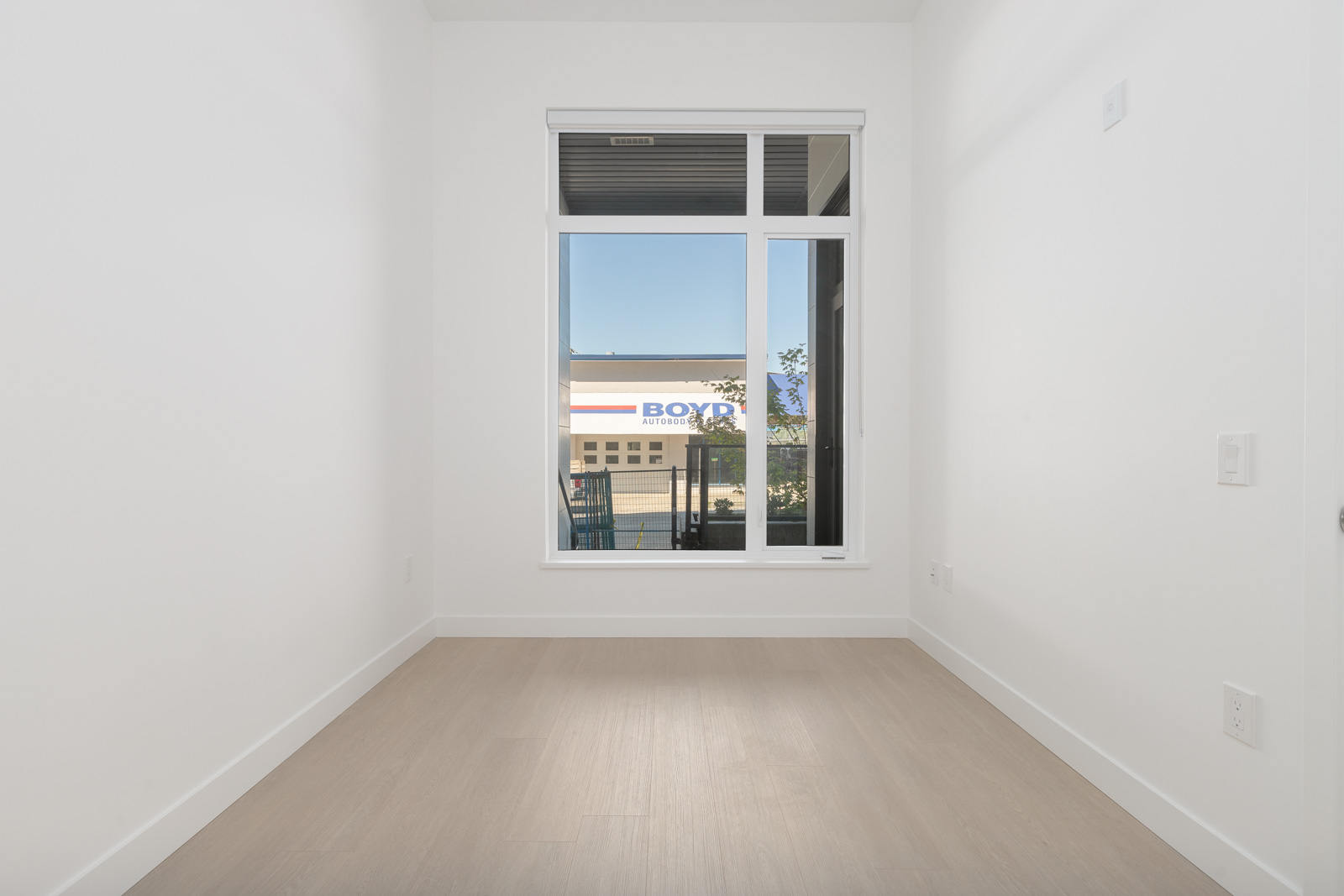Small, empty room with light wood flooring, white walls, and a tall window showing a building and tree outside.