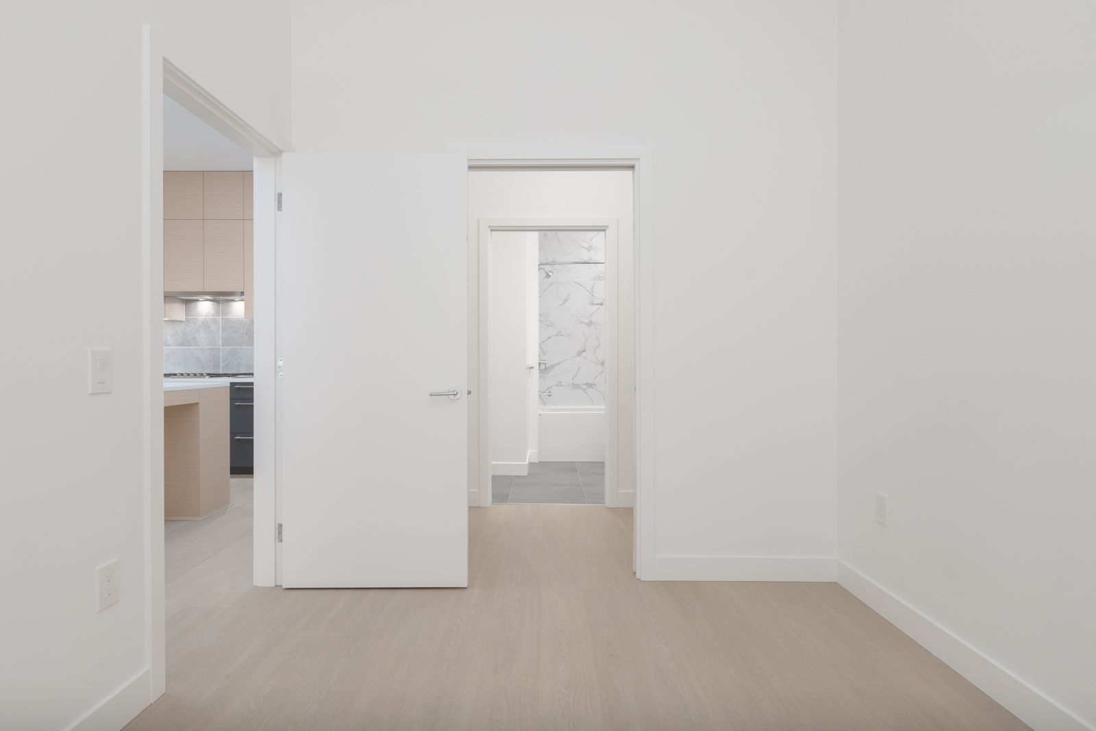 A view of an empty room with light wood flooring, an open door leading to a kitchen on the left, and a doorway straight ahead showing a bathroom with a bathtub.