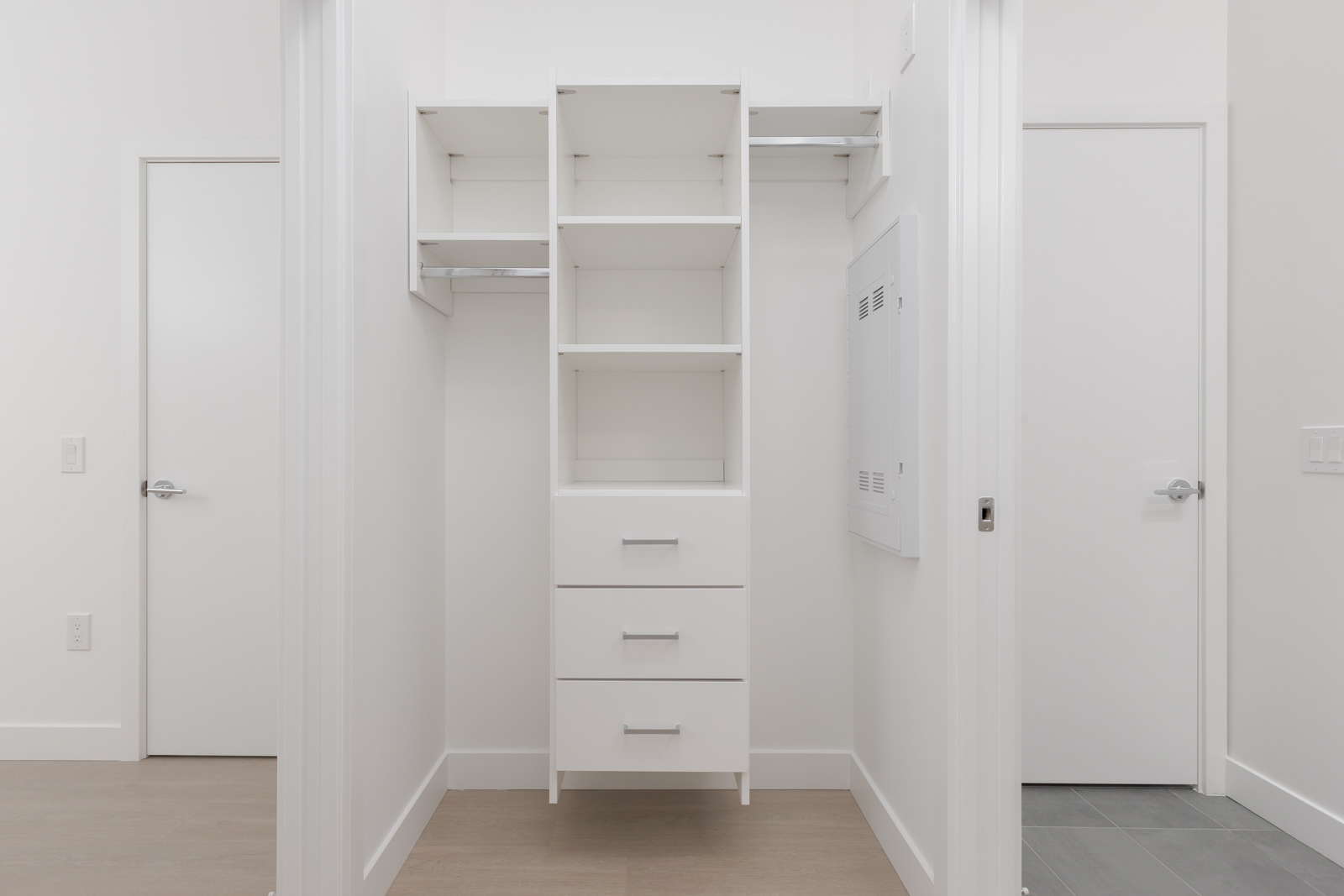 Empty walk-in closet with white shelves, drawers, and hanging rods, viewed from the doorway. The closet has neutral-colored walls and flooring.