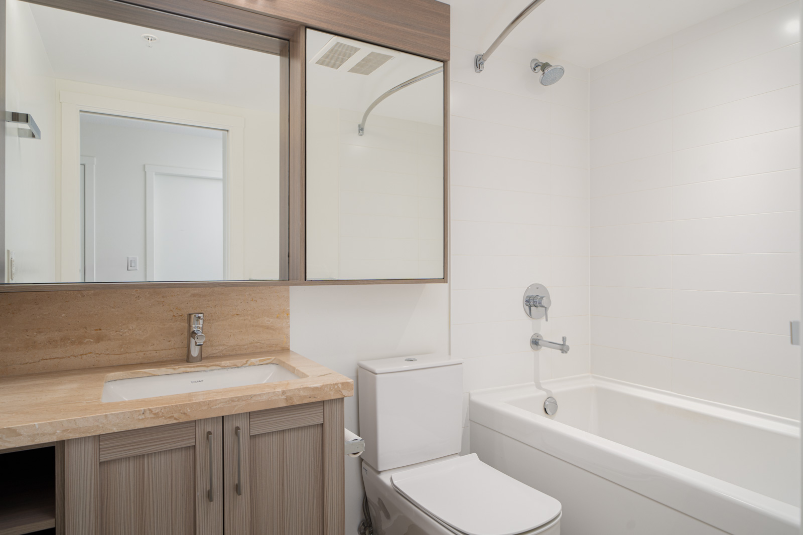 Modern bathroom with a beige countertop, sink, toilet, bathtub, showerhead, large mirror, and wood cabinetry. White tiles cover the walls around the tub.