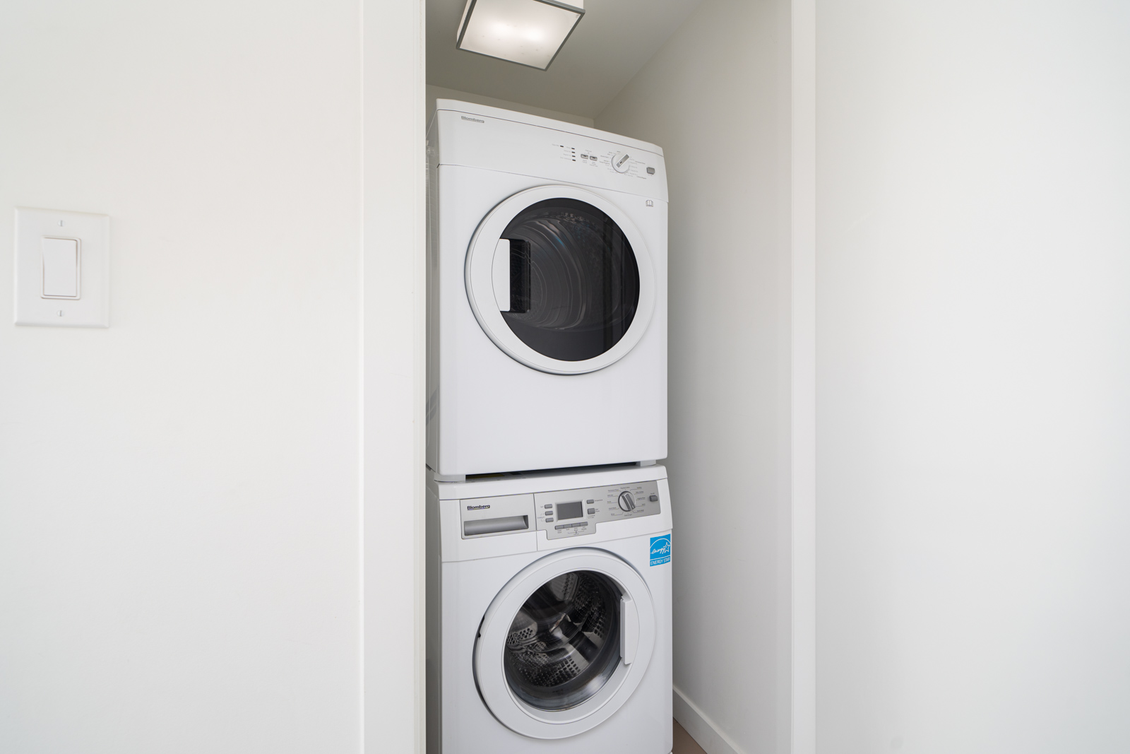 A white stacked washer and dryer unit in a small, recessed laundry nook with white walls and a ceiling light above.